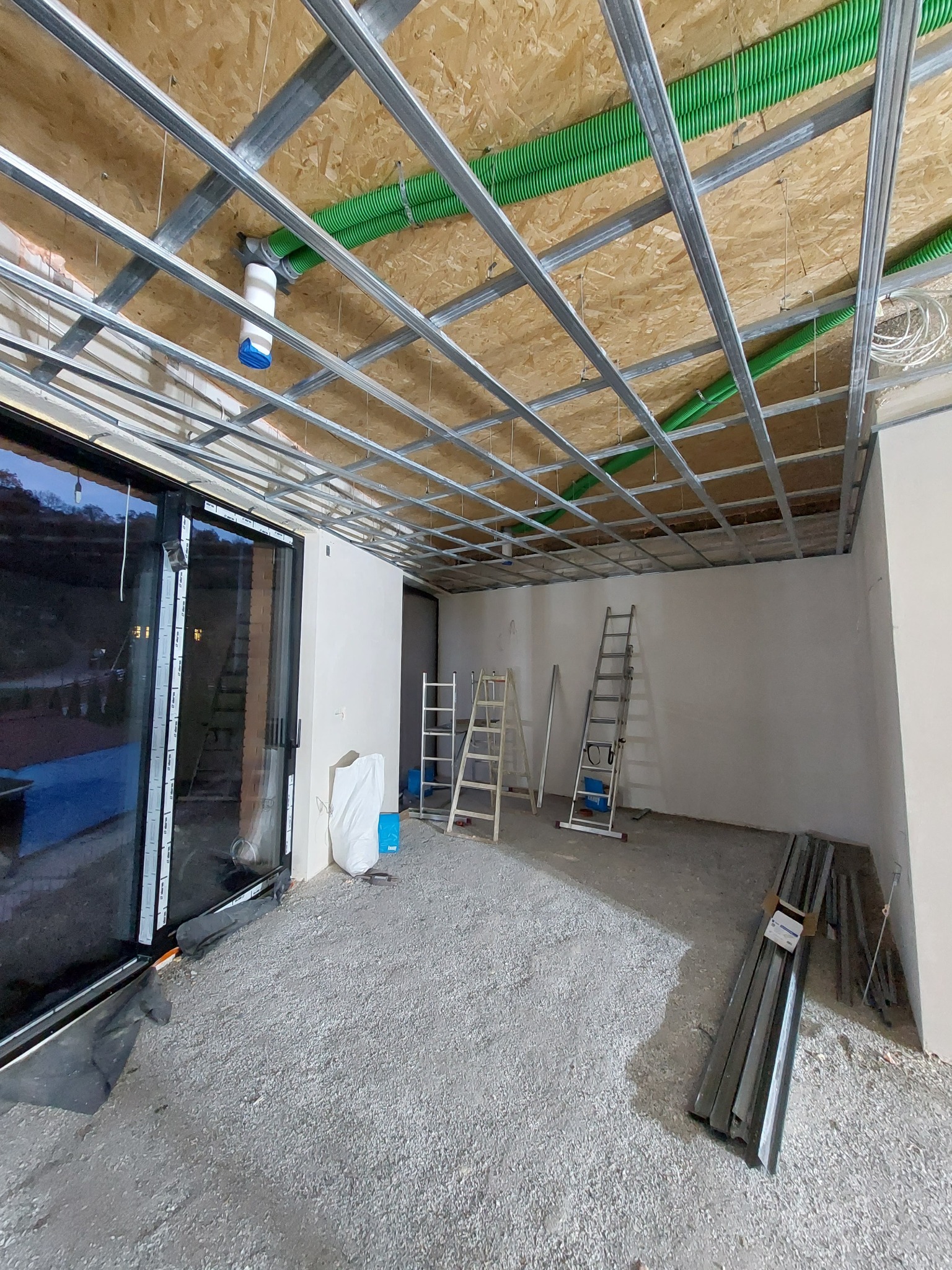 Montaż metalowego stelażu sufitu podwieszanego z widocznymi rurami instalacji elektrycznej w kolorze zielonym, drabiny oparta o ścianę, surowy stan wykończenia, widok z dołu.