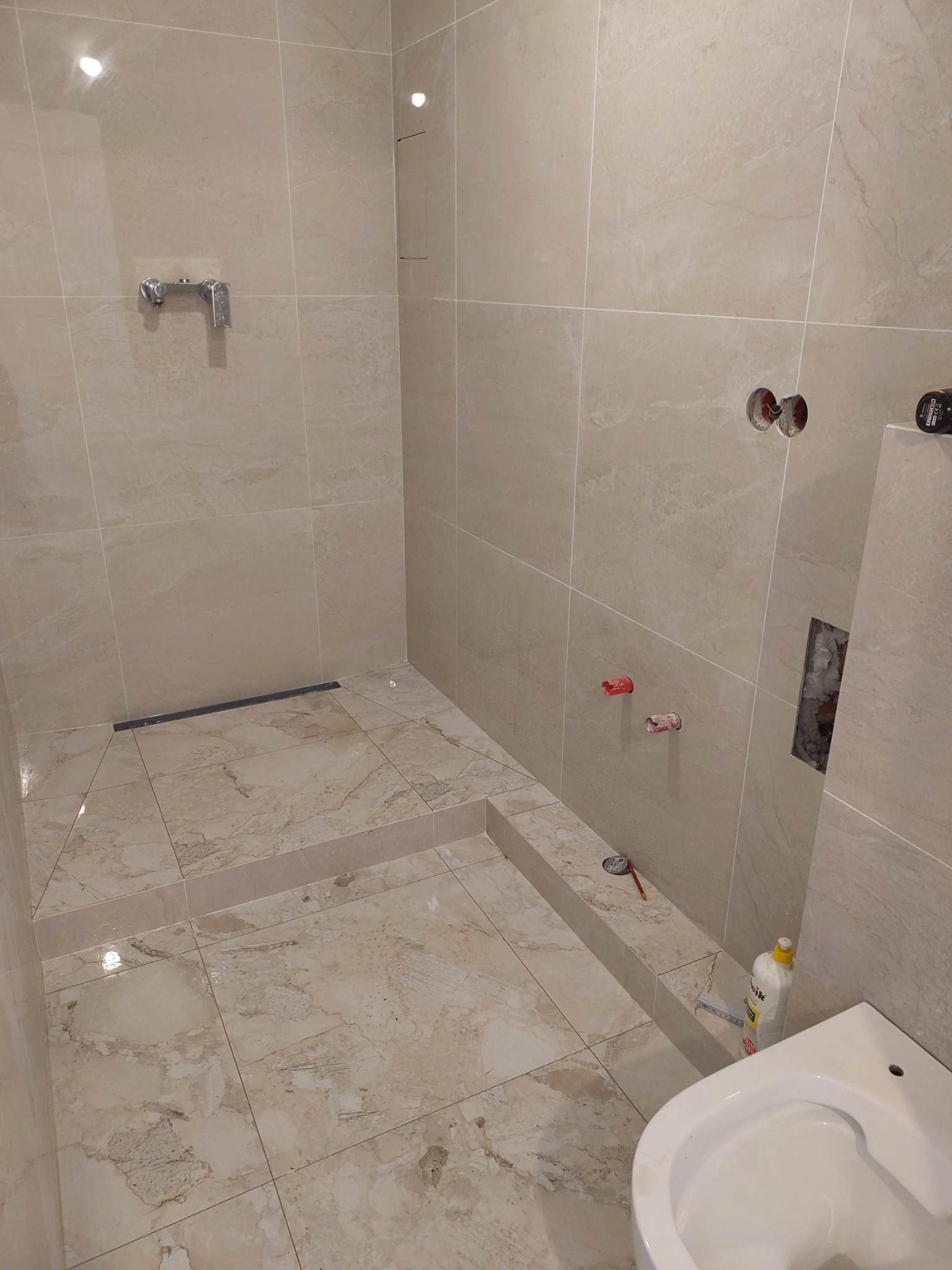 Łazienka w trakcie wykańczania: beżowe płytki podłogowe i ścienne z marmurkowym wzorem, zamontowany odpływ liniowy w strefie prysznica, widoczne przyłącza wodne i kanalizacyjne, częściowo...