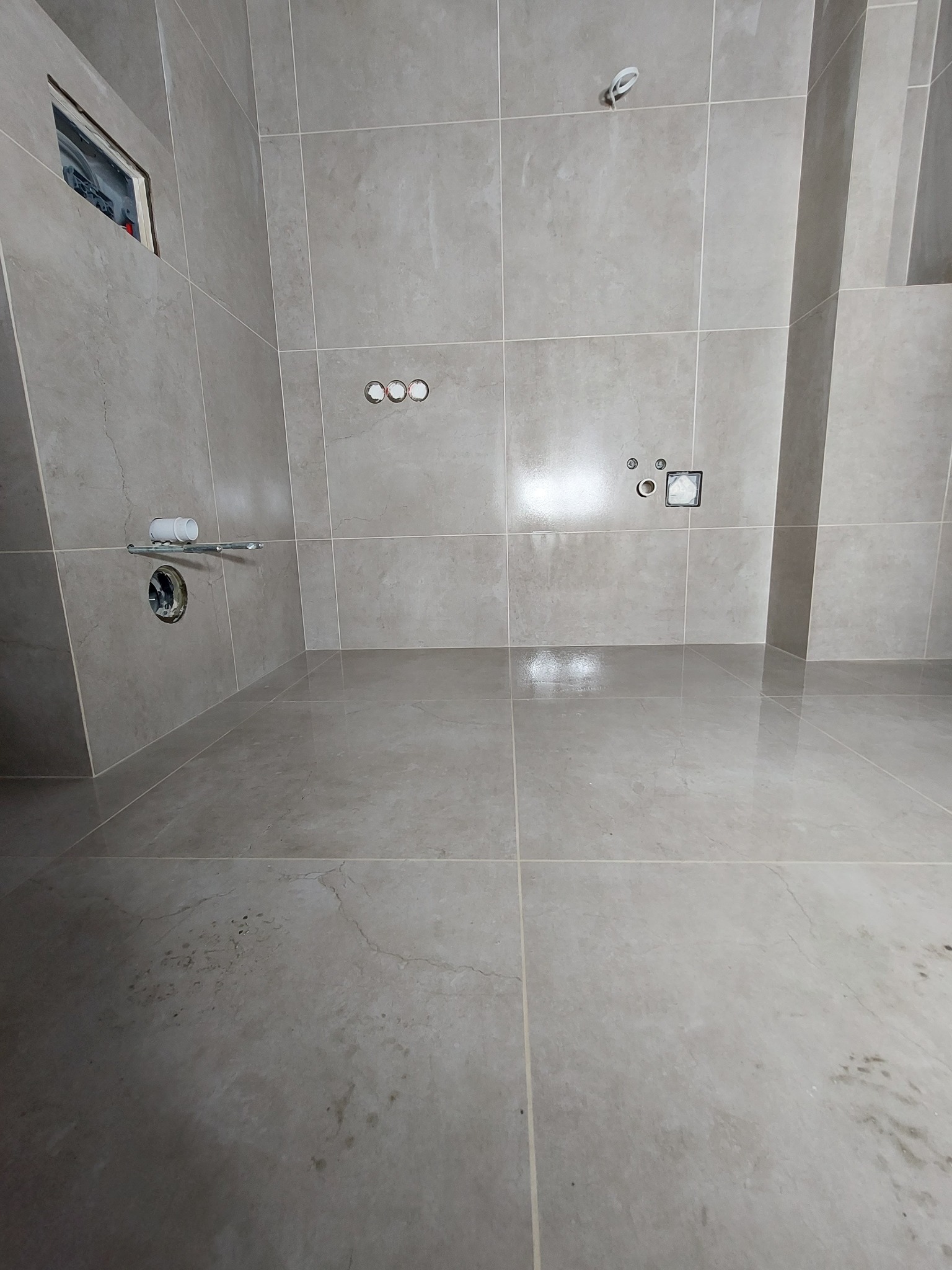 Łazienka w trakcie remontu, widoczne szare płytki na ścianach i podłodze, przygotowane otwory na instalacje, elementy instalacji sanitarnej wystające ze ściany, oraz otwór rewizyjny.