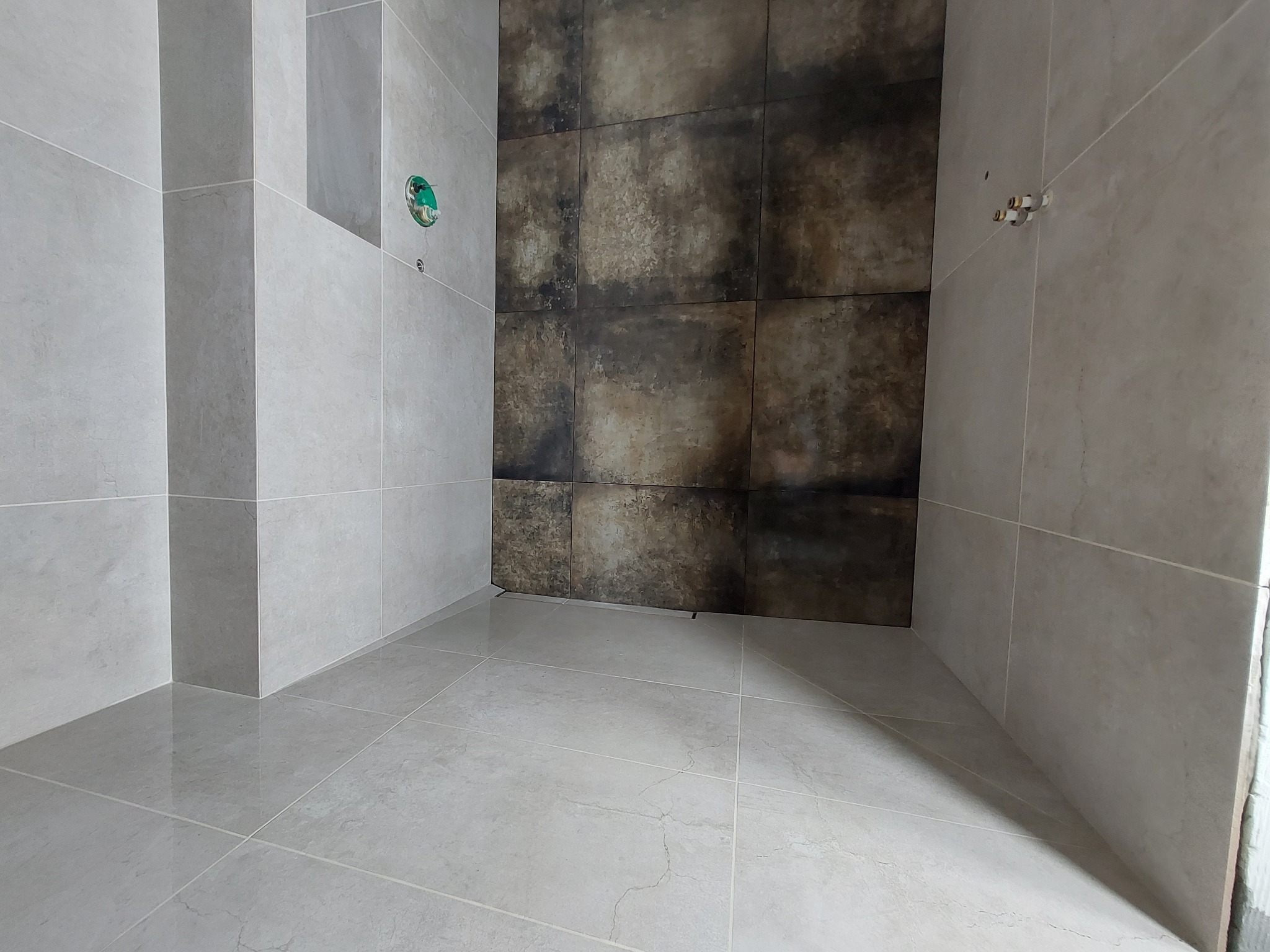 Wykończona łazienka z szarymi płytkami na ścianach i podłodze, akcentem jest ściana z ciemnymi płytkami imitującymi rdzę, widoczne przyłącza wodne.