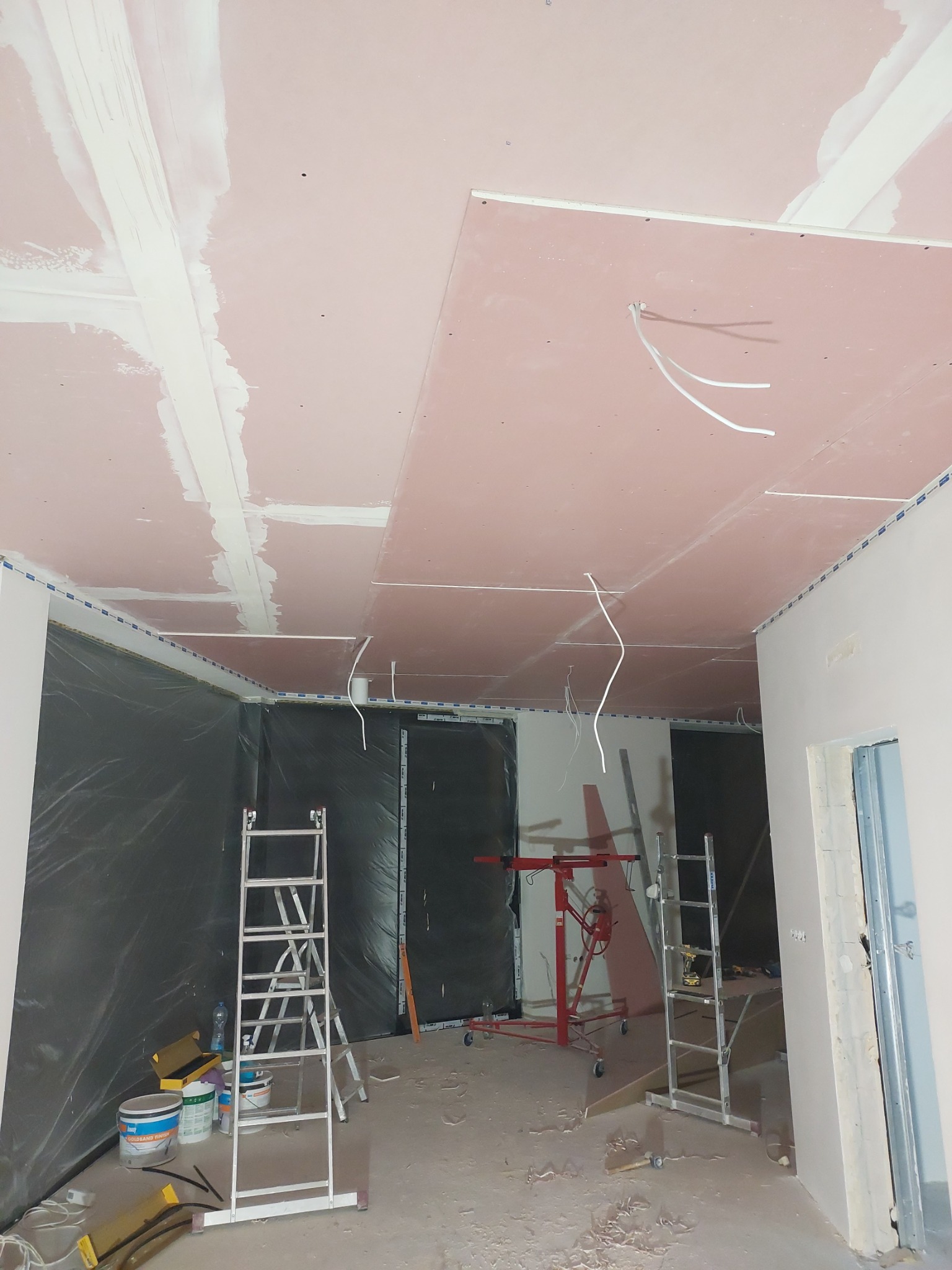 Montaż sufitu z płyt karton-gips: widoczne płyty w kolorze różowym, częściowo zaszpachlowane łączenia, wystające przewody elektryczne, drabiny rozstawione w pomieszczeniu.