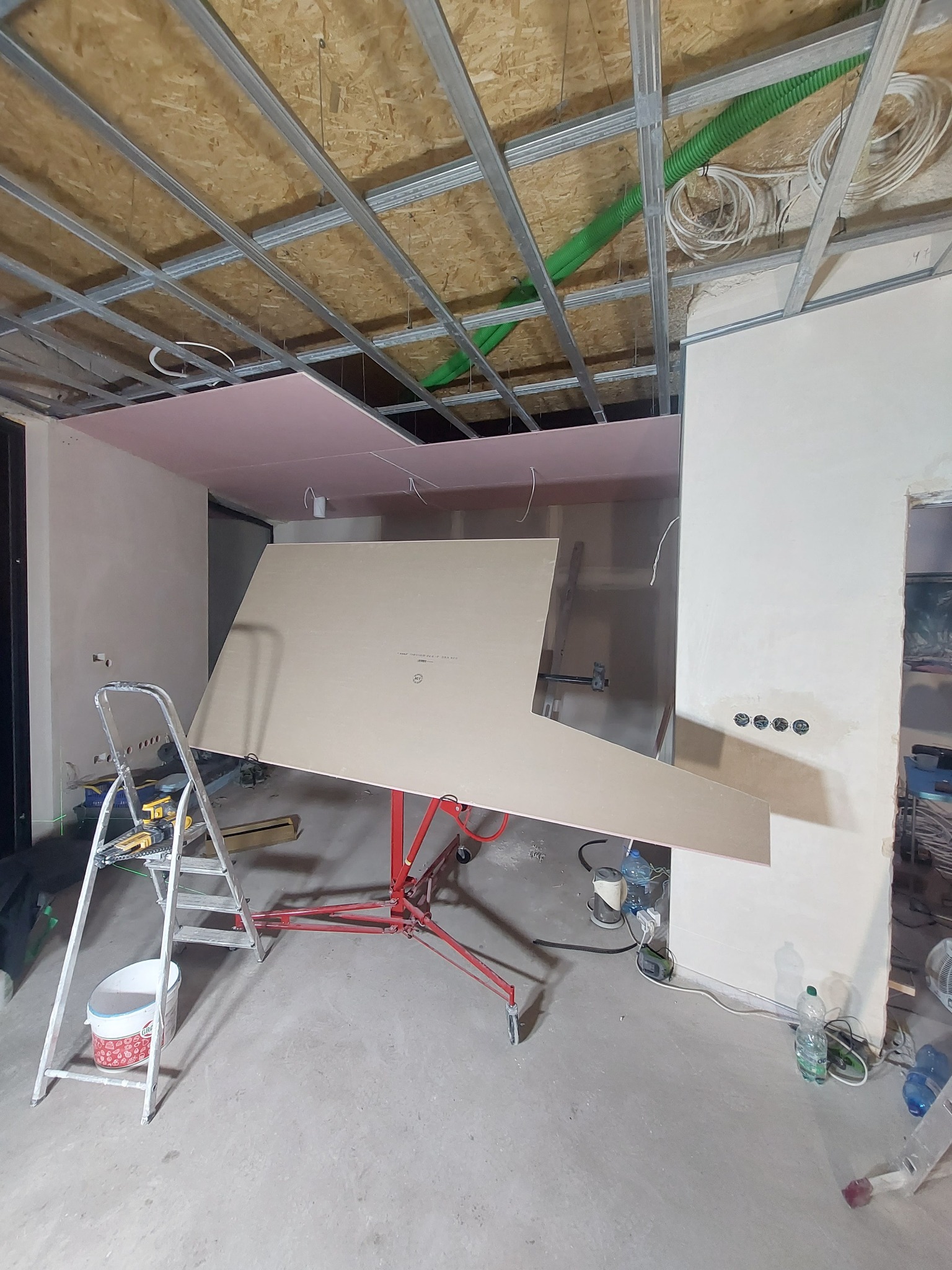 Montaż płyty gipsowo-kartonowej na podnośniku w trakcie remontu, widoczne profile sufitowe, drabina z narzędziami i instalacje elektryczne.