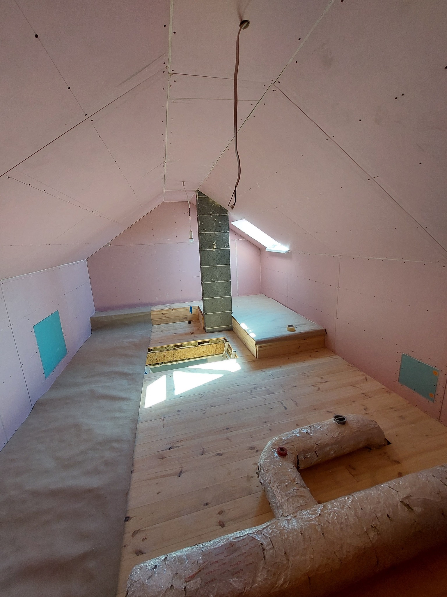 Poddasze w trakcie adaptacji: ściany i skosy obłożone różową płytą karton-gips, drewniana podłoga częściowo zakryta papierem ochronnym, widoczny komin z szarej cegły i izolowane rury wentylacyjne.