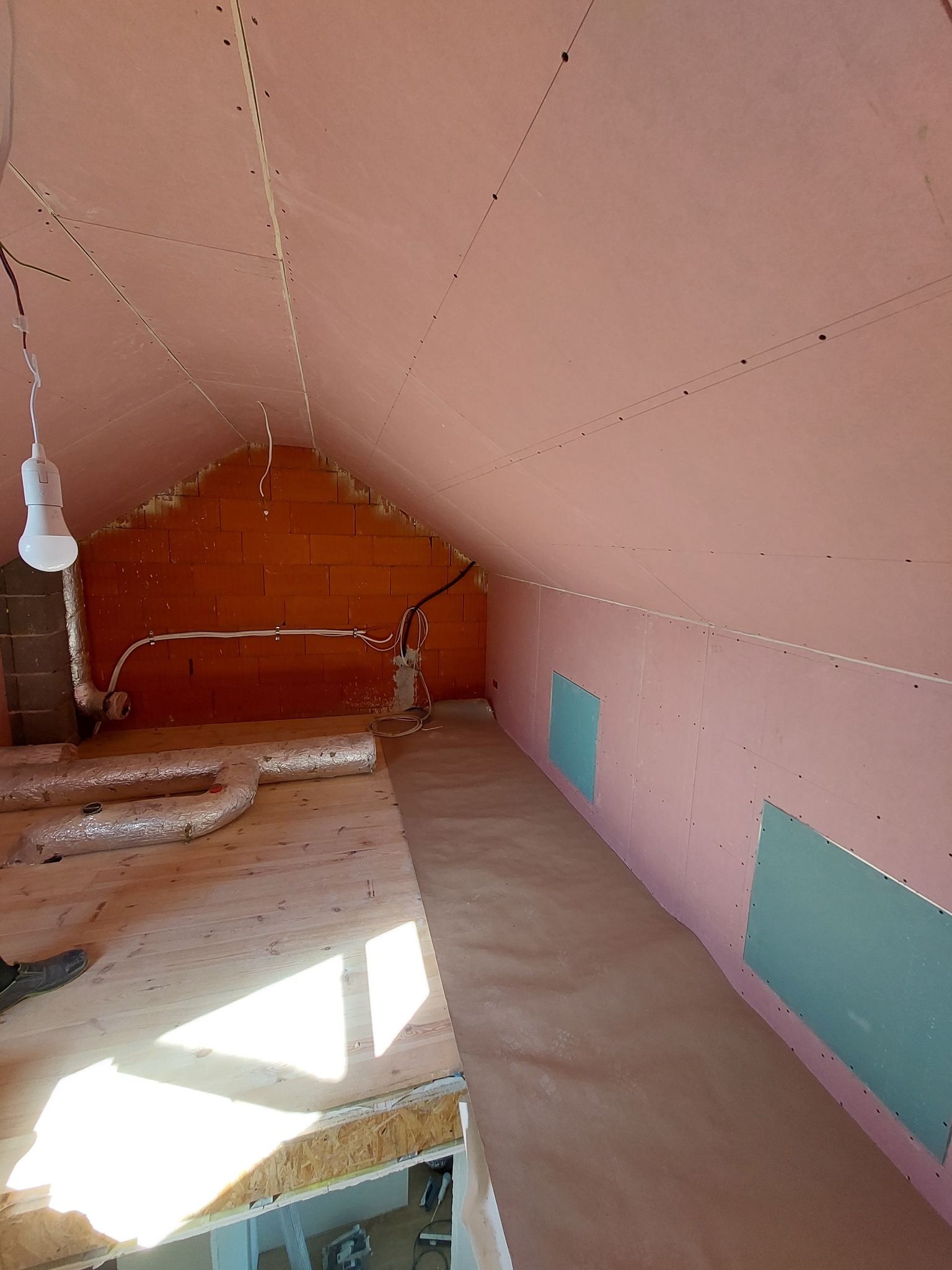 Poddasze w trakcie wykańczania: ściany i skośny sufit obłożone różową płytą karton-gips, widoczne łączenia płyt, fragment surowej ściany z cegły, instalacja elektryczna i rury wentylacyjne, podłoga...