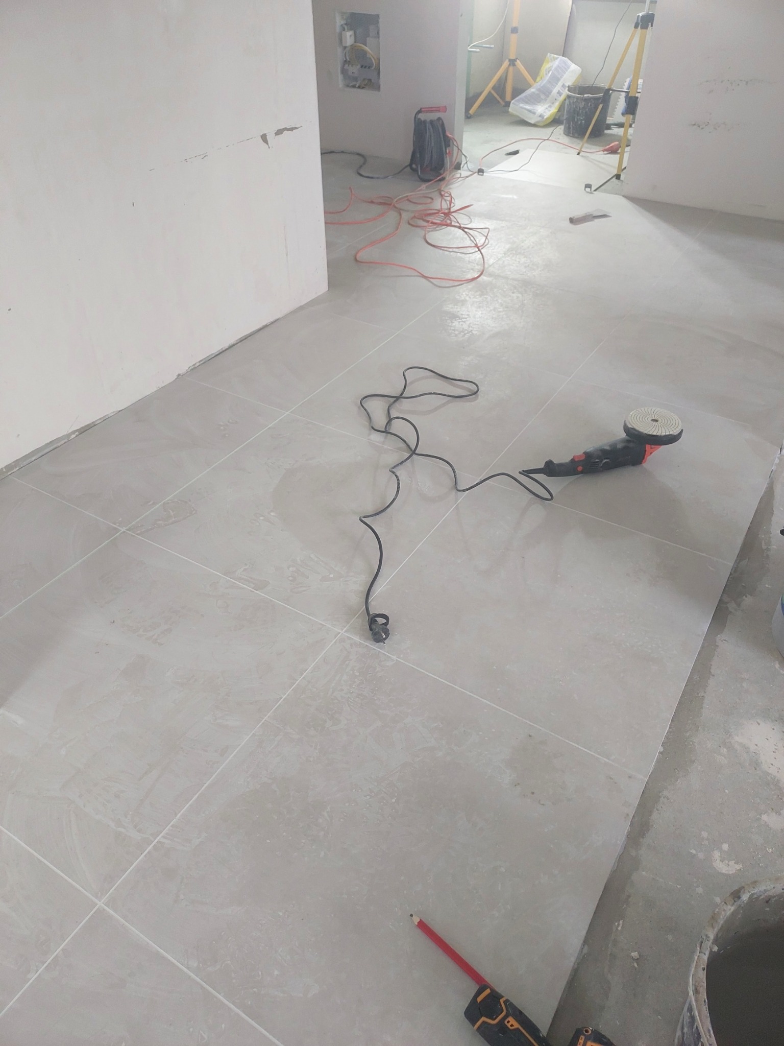 Świeżo położone, jasnoszare płytki podłogowe, częściowo mokre, z widoczną polerką do płytek, kablem zasilającym, ołówkiem i wiadrem w rogu pomieszczenia w trakcie prac wykończeniowych.