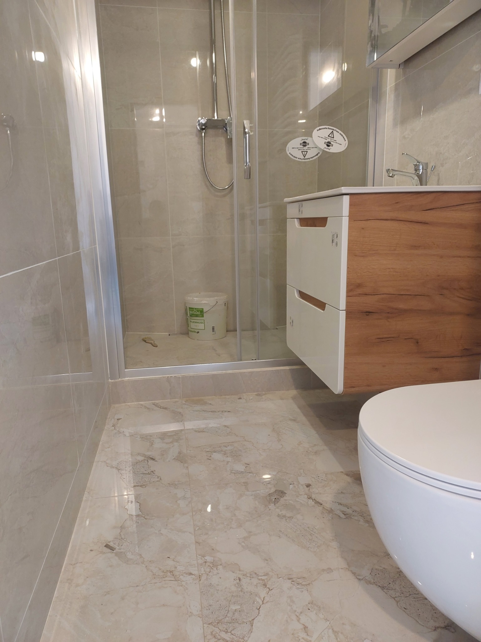 Wykończona łazienka z beżowymi płytkami imitującymi marmur na podłodze i ścianach, kabina prysznicowa ze szklanymi drzwiami, wisząca szafka pod umywalką z drewnianym frontem i białą obudową...