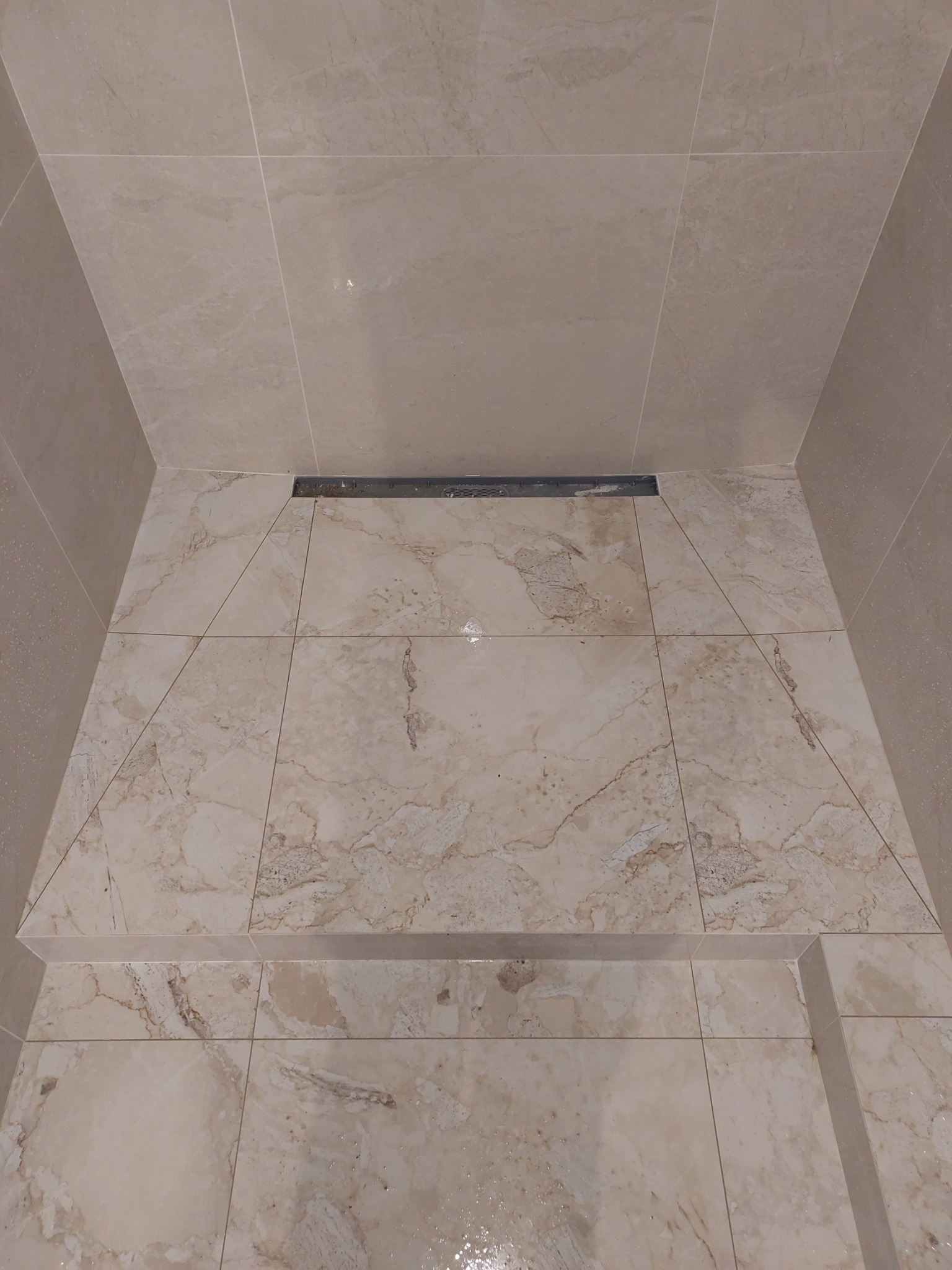 Wykończony brodzik z beżowych płytek imitujących marmur, z odpływem liniowym przy ścianie, widoczne fugi i delikatne załamania światła.