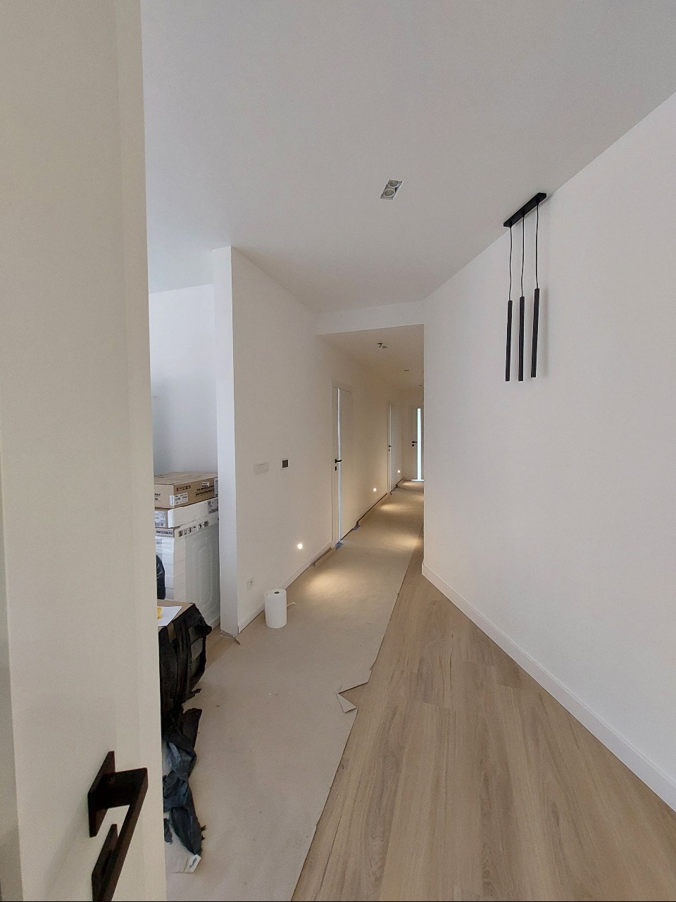 Korytarz w trakcie prac wykończeniowych: podłoga częściowo zabezpieczona kartonem, białe ściany z zamontowanym oświetleniem punktowym i wiszącą lampą, widoczne materiały budowlane w tle.