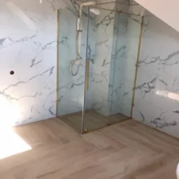 Wykończona łazienka na poddaszu z kabiną prysznicową ze złotymi profilami, marmurowe płytki na ścianach i drewnopodobna podłoga ułożona w jodełkę.