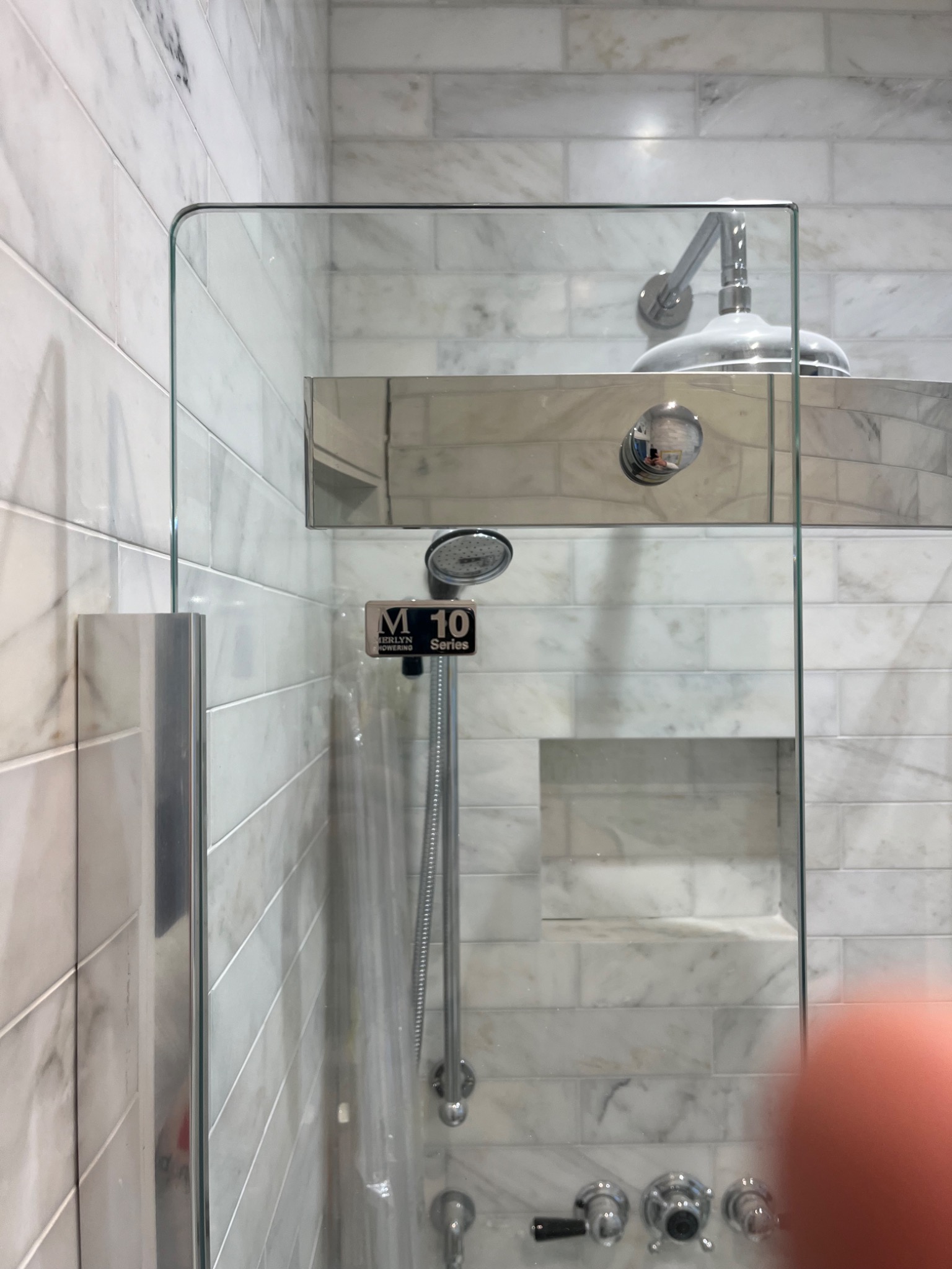 Szklana kabina prysznicowa z chromowaną armaturą na tle marmurowych płytek ułożonych w cegiełkę, widoczna etykieta 'M 10 Series' na słuchawce prysznicowej.