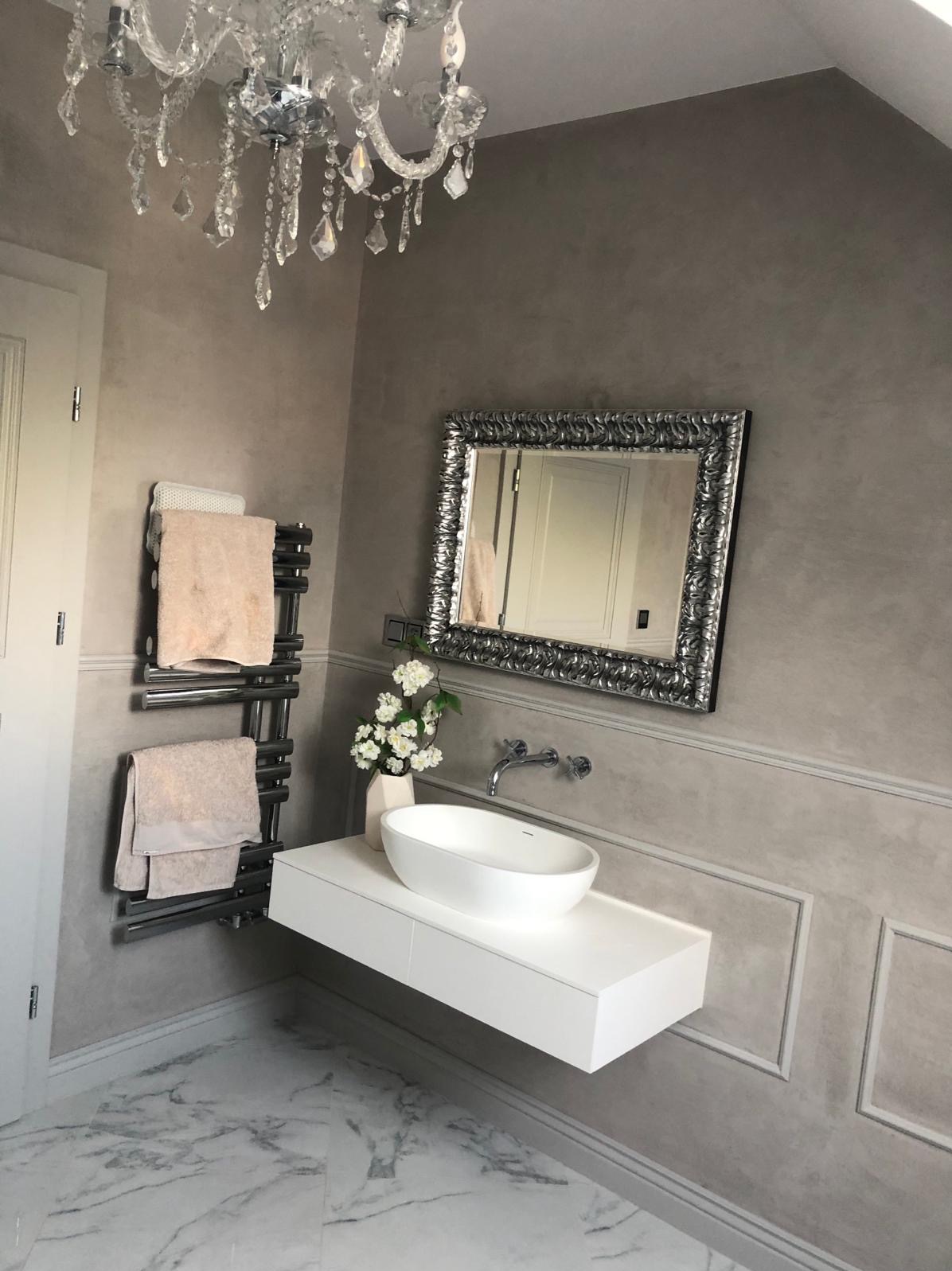 Elegancka łazienka z szarymi ścianami, marmurową podłogą i kryształowym żyrandolem. Umywalka nablatowa o owalnym kształcie na białej szafce wiszącej, lustro w ozdobnej srebrnej ramie, grzejnik...