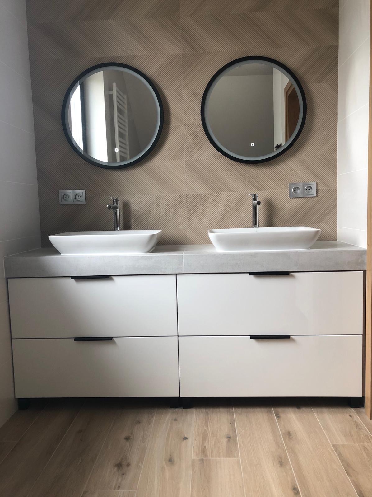Nowoczesna łazienka z dwoma okrągłymi lustrami z podświetleniem LED, dwiema białymi umywalkami nablatowymi na szarym blacie i białymi szafkami z czarnymi uchwytami, na ścianie beżowe płytki ułożone...