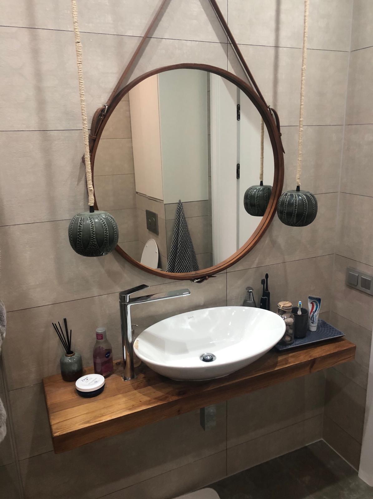 Nowoczesna łazienka z okrągłym lustrem na skórzanym pasku, wiszącym nad nablatową umywalką na drewnianym blacie, z dodatkami w odcieniach szarości i bieli.