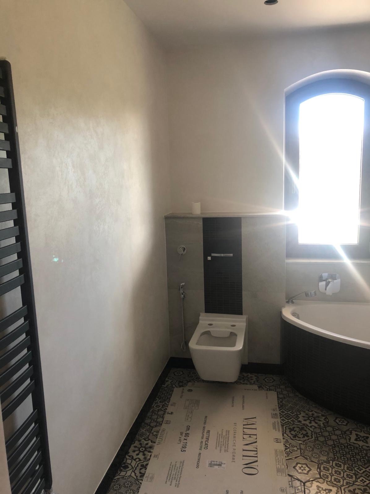 Wykończona łazienka z beżową ścianą, czarnym grzejnikiem drabinkowym, toaletą, wanną z czarną obudową i oknem łukowym. Na podłodze leżą kartony z napisem VALENTINO.