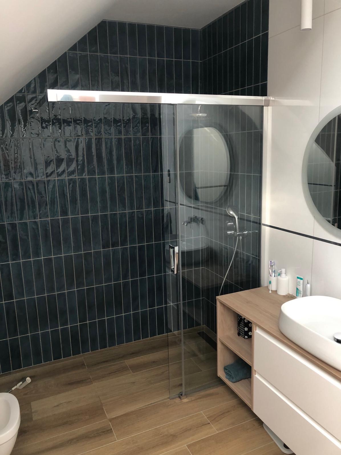 Nowoczesna łazienka na poddaszu z kabiną prysznicową ze szklanymi drzwiami przesuwnymi, ciemnymi, pionowo ułożonymi płytkami na ścianie pod skosem, drewnopodobną podłogą i białą umywalką nablatową...