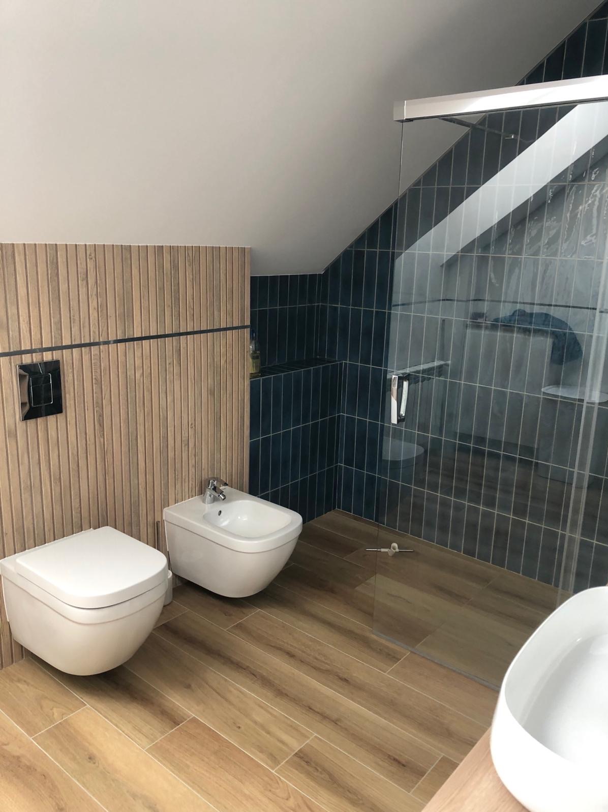 Nowoczesna łazienka na poddaszu z drewnopodobną glazurą pionową, bidetem i toaletą podwieszaną oraz kabiną prysznicową z ciemnymi, wąskimi płytkami.
