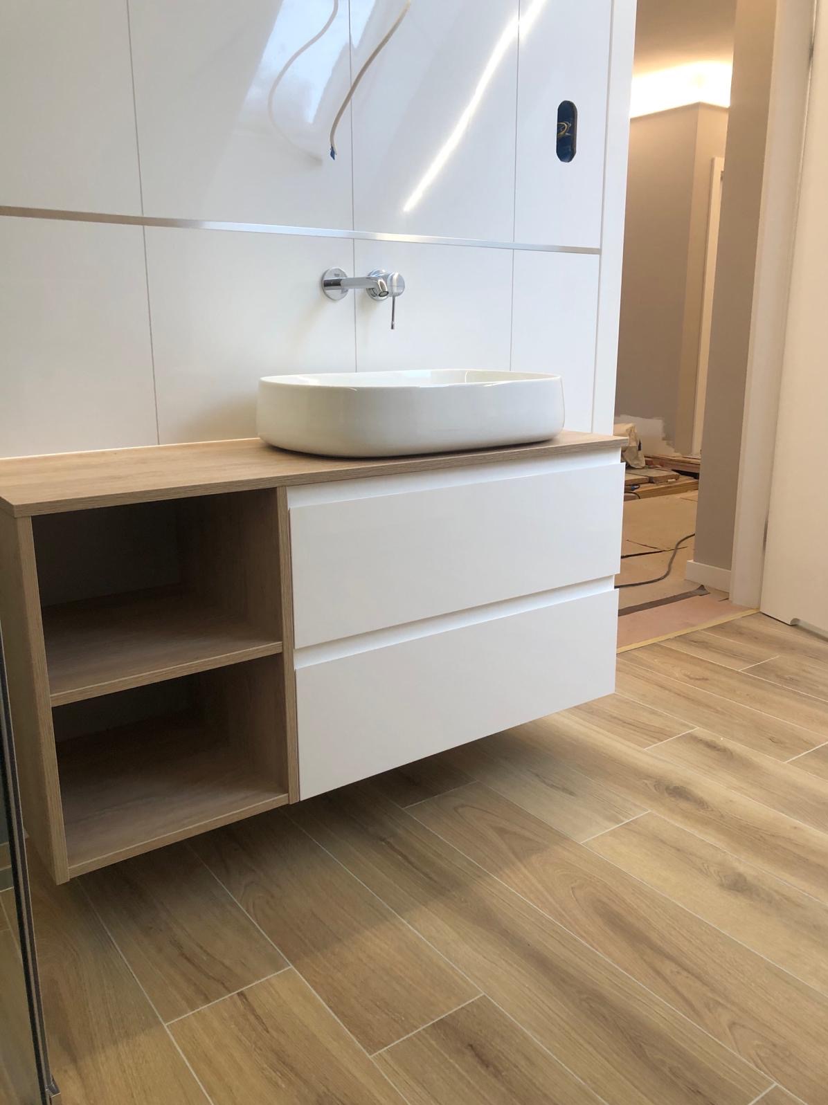 Nowoczesna łazienka z owalną umywalką nablatową, szafką z szufladami i półkami oraz płytkami imitującymi drewno na podłodze, widoczne niedokończone prace wykończeniowe w tle.