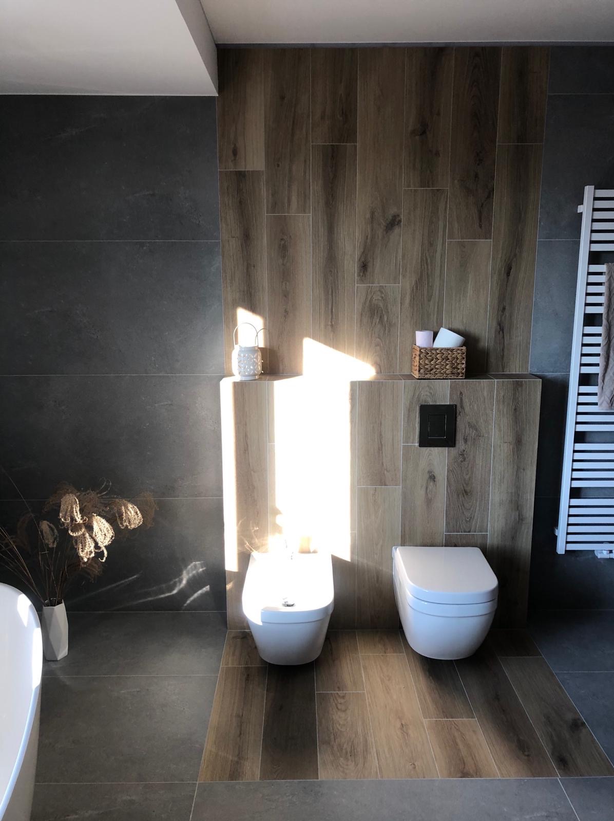 Nowoczesna łazienka z szarymi i drewnopodobnymi płytkami, wisząca toaleta i bidet, grzejnik drabinkowy, dekoracje w postaci lampionu i suszonych traw, oświetlona promieniami słońca.