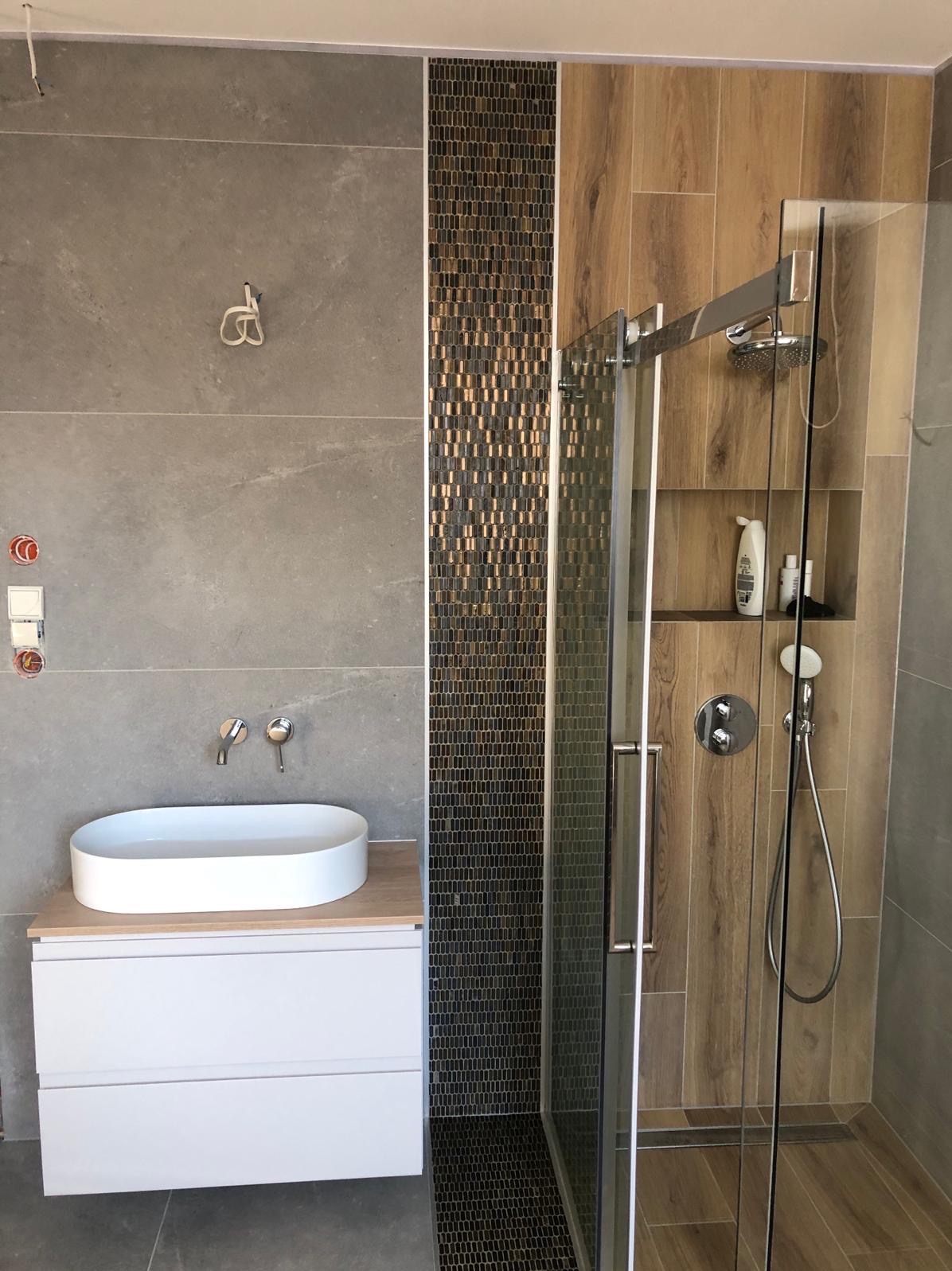 Nowoczesna łazienka z kabiną prysznicową ze szklanymi drzwiami, umywalką nablatową na białej szafce, szarą płytką ścienną, drewnopodobną okładziną prysznica i pionowym pasem mozaiki w odcieniach...