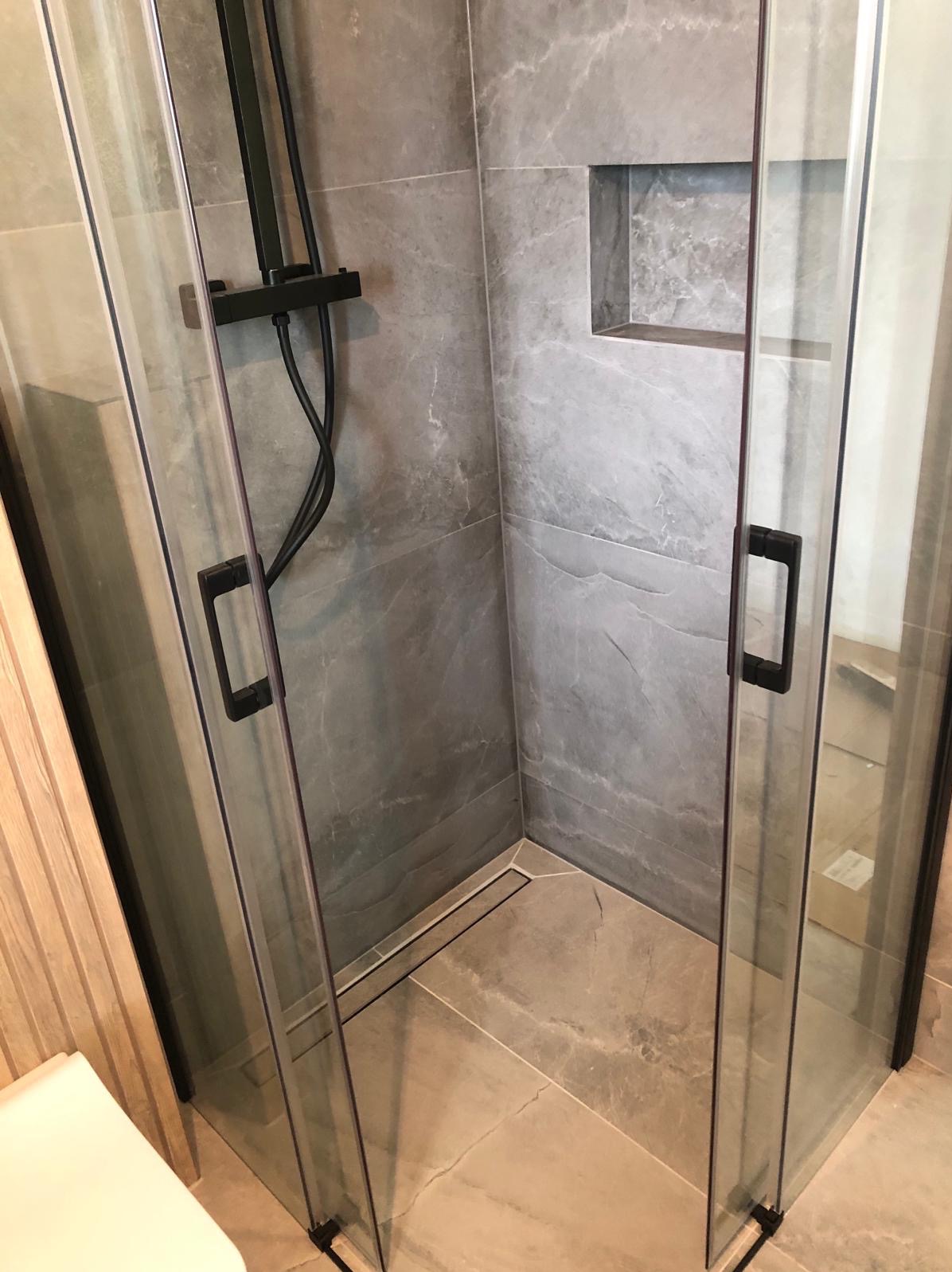 Nowoczesna kabina prysznicowa z czarnymi elementami, szarymi płytkami imitującymi marmur i odpływem liniowym w podłodze.