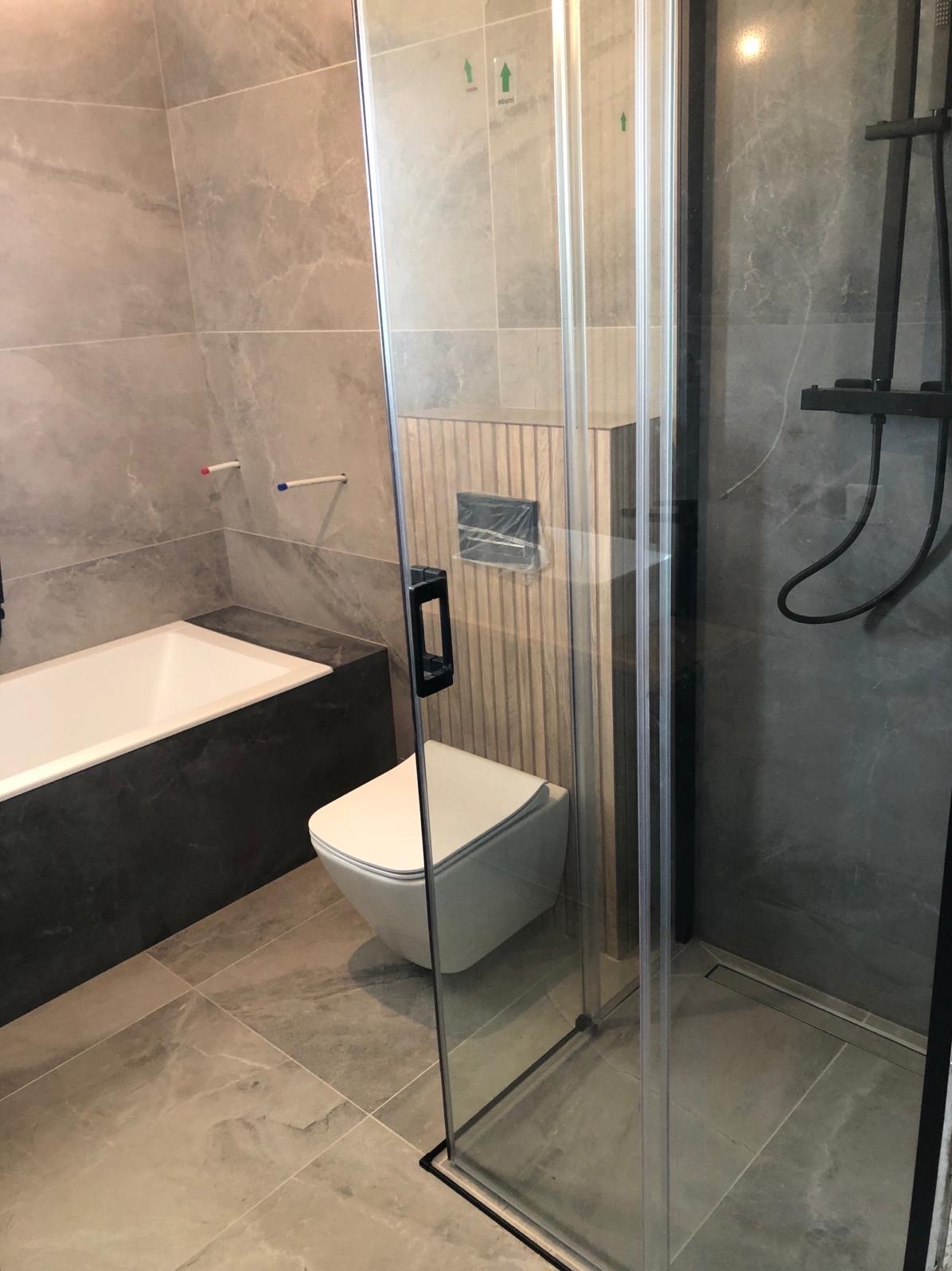 Nowoczesna łazienka w szarościach z kabiną prysznicową typu walk-in z czarnymi profilami, podwieszaną toaletą i wanną obudowaną ciemnym kamieniem.