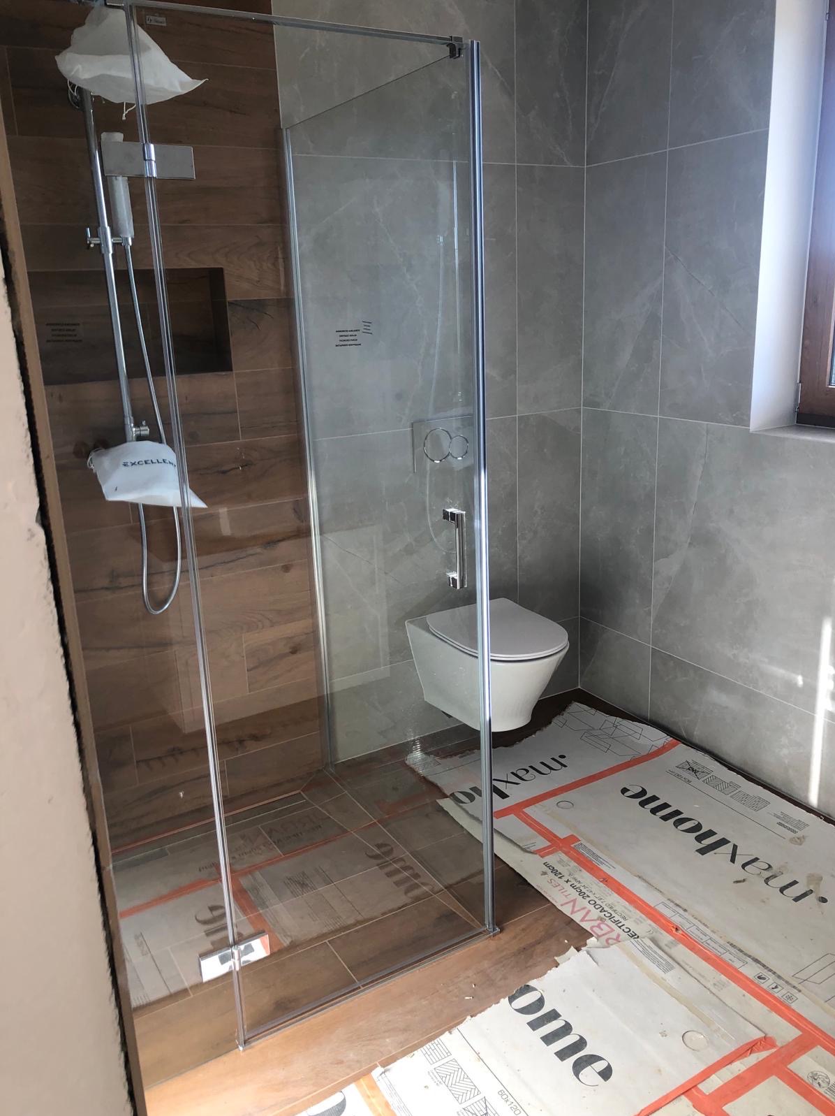 Nowoczesna łazienka z kabiną prysznicową typu walk-in, drewnopodobnymi płytkami na ścianie prysznica i szarymi płytkami na pozostałych ścianach, wisząca toaleta oraz folia ochronna na podłodze.