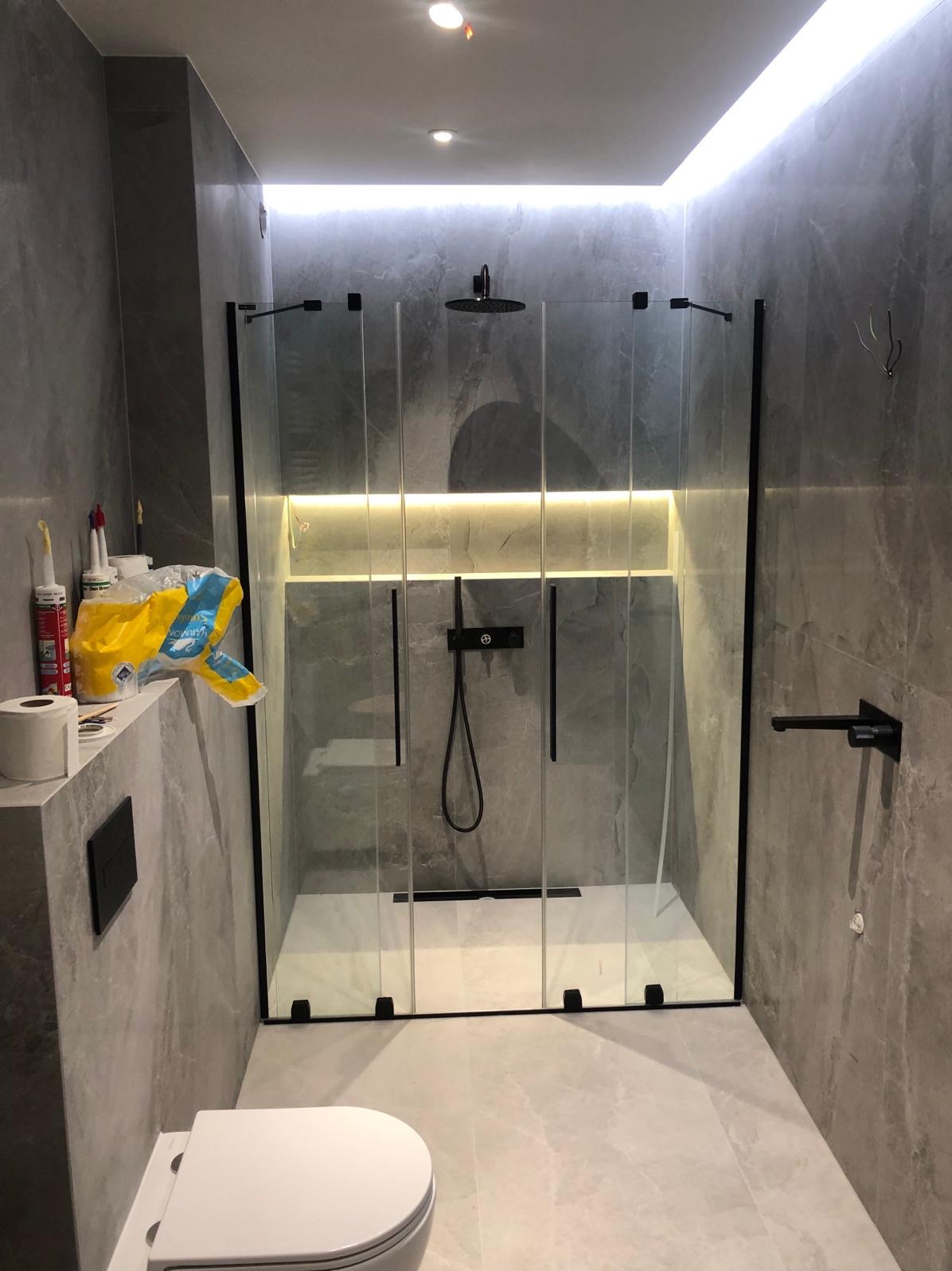 Nowoczesna łazienka z kabiną prysznicową typu walk-in z czarnymi ramami, wykończona płytkami imitującymi marmur, z podświetleniem LED w niszy prysznicowej i widocznymi akcesoriami montażowymi...