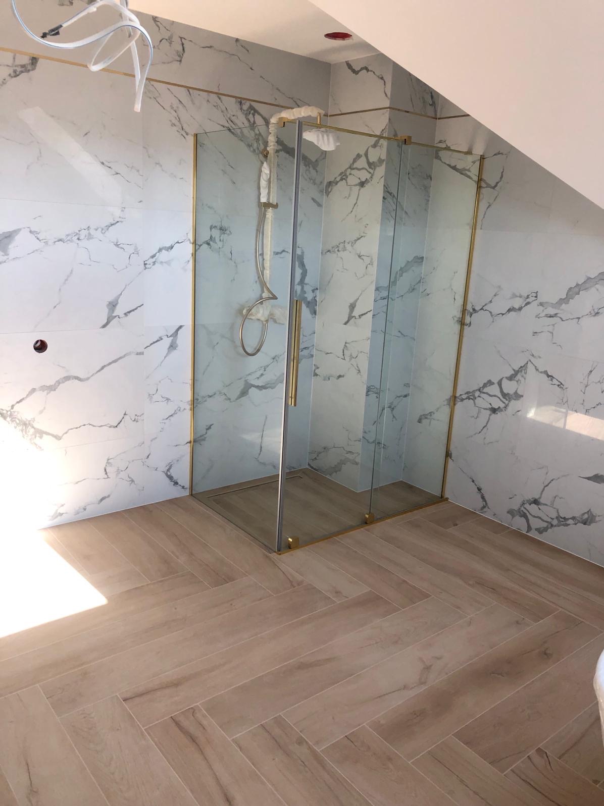 Wykończona łazienka na poddaszu z kabiną prysznicową ze złotymi profilami, marmurowe płytki na ścianach i drewnopodobna podłoga ułożona w jodełkę.