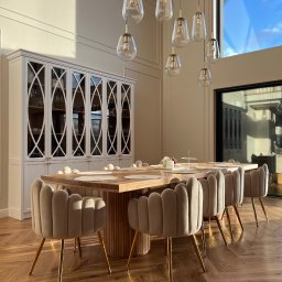 Rem-Mar - Jadalnia z dębowym stołem na okrągłej ryflowanej podstawie, otoczonym beżowymi krzesłami na złotych nogach, na parkiecie ułożonym w jodełkę, z białym kredensem w tle i designerskimi lampami wiszącymi.