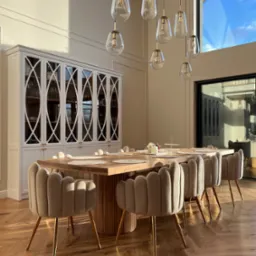 Jadalnia z dębowym stołem na okrągłej ryflowanej podstawie, otoczonym beżowymi krzesłami na złotych nogach, na parkiecie ułożonym w jodełkę, z białym kredensem w tle i designerskimi lampami wiszącymi.
