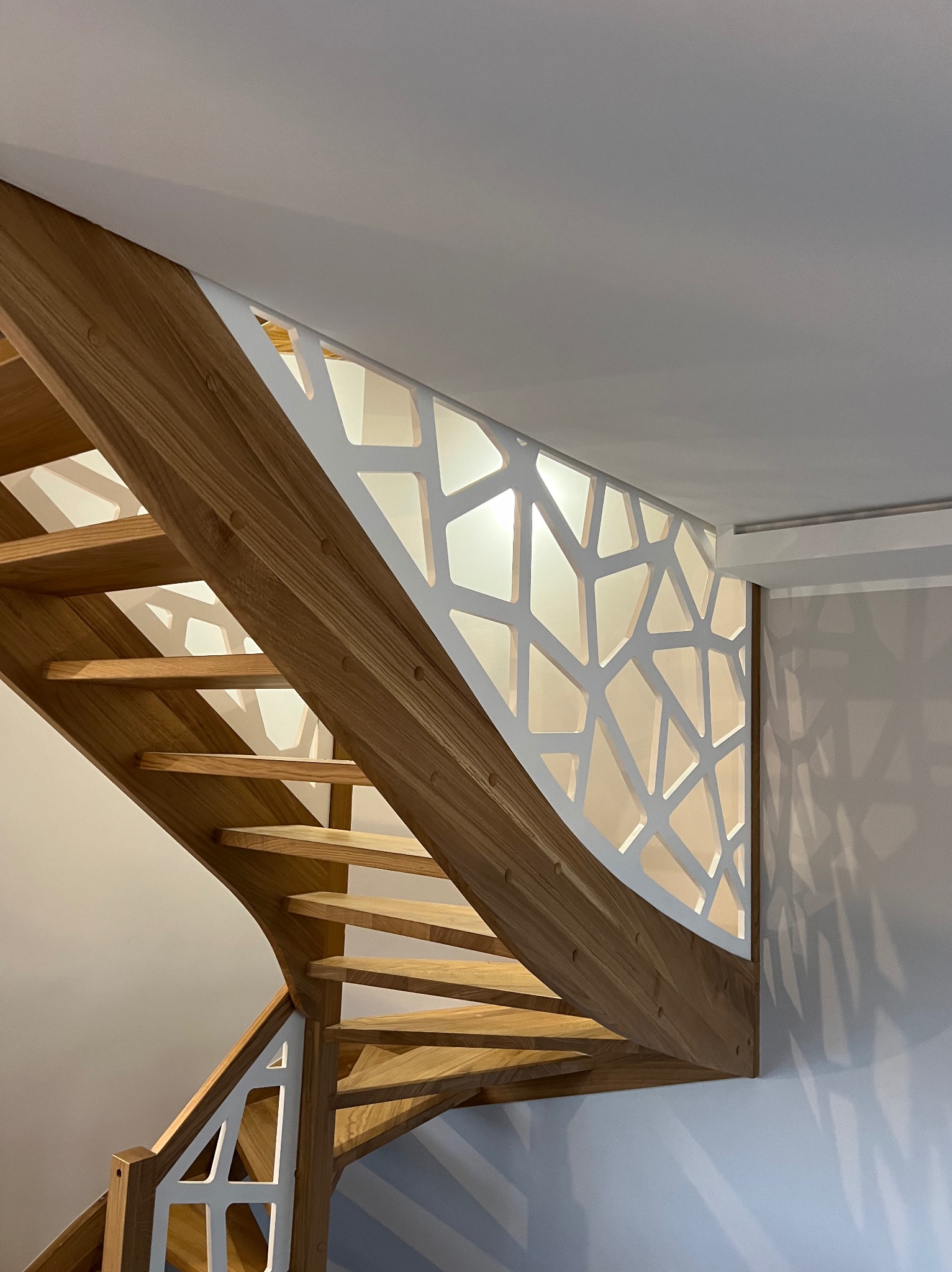 Nowoczesne schody z jasnego drewna z ażurową, białą balustradą o geometrycznym wzorze, oświetlone od spodu, widoczne z dołu.