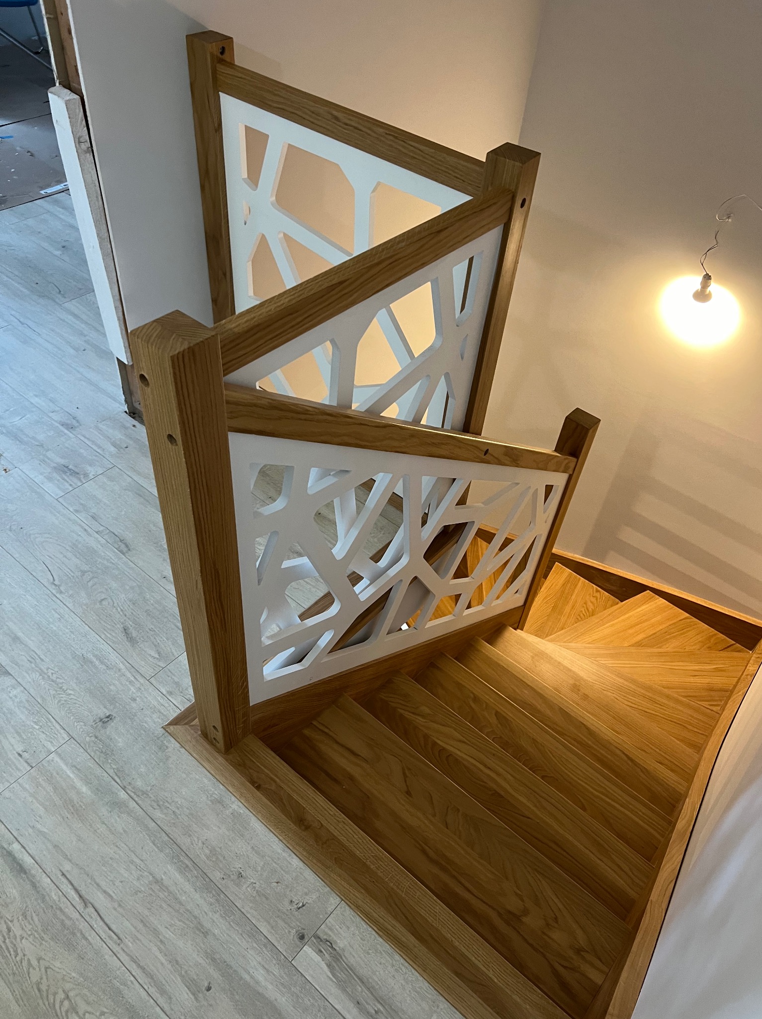 Jasne, drewniane schody z geometryczną, ażurową balustradą w kolorze białym, widziane z góry, z delikatnym oświetleniem.