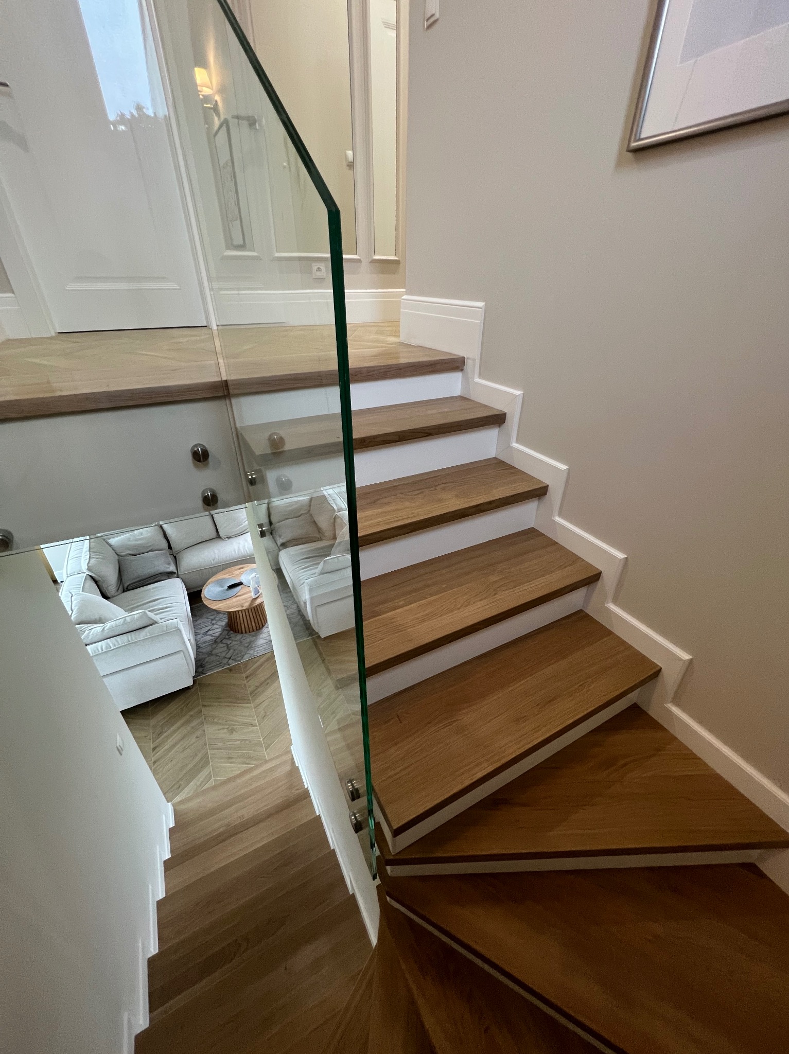 Nowoczesne schody drewniane z przeszkloną balustradą, widok z góry na salon z jasną sofą.