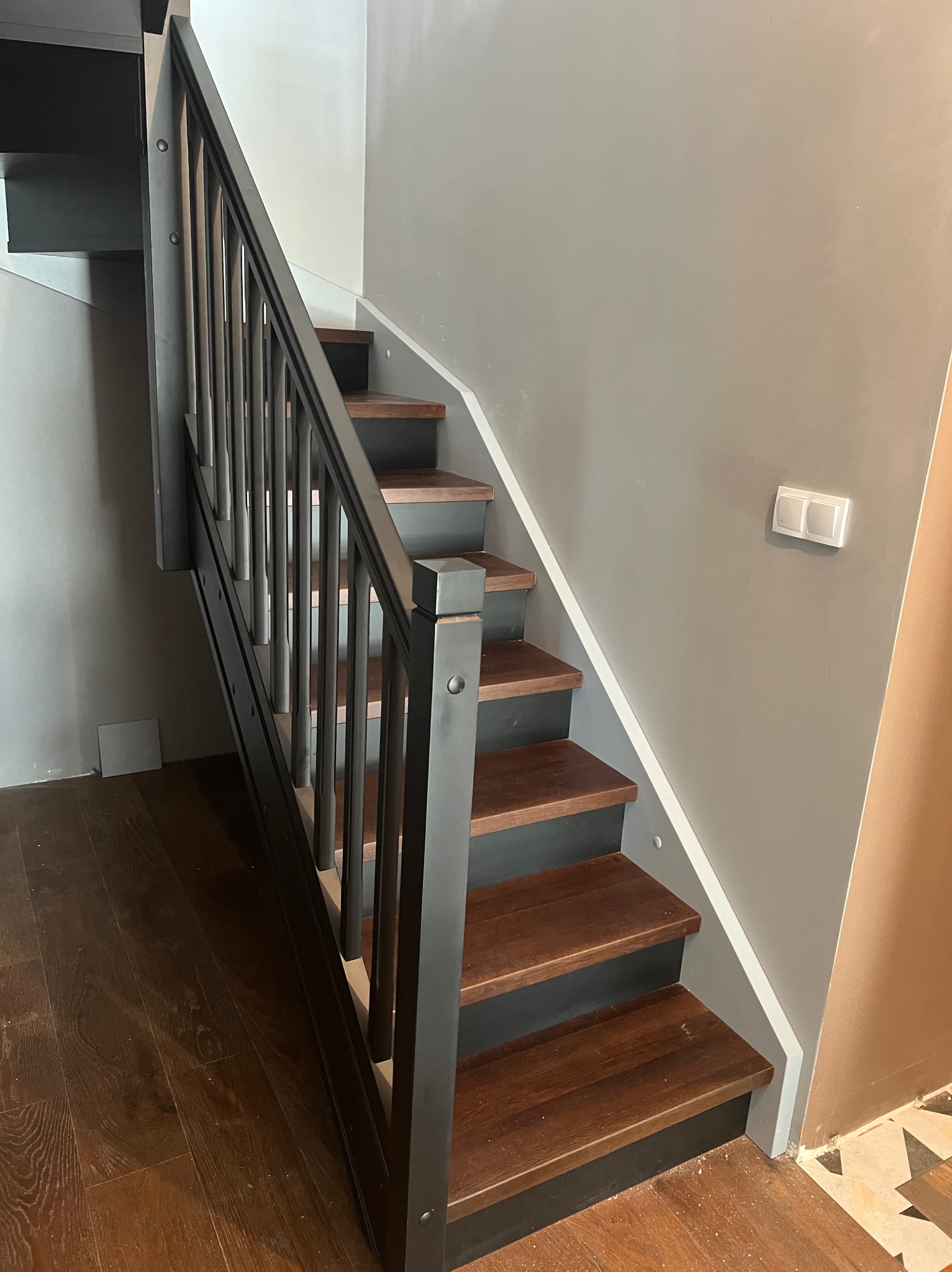 Nowoczesne schody z drewnianymi stopniami i czarną metalową balustradą w minimalistycznym wnętrzu z szarą ścianą i drewnianą podłogą.