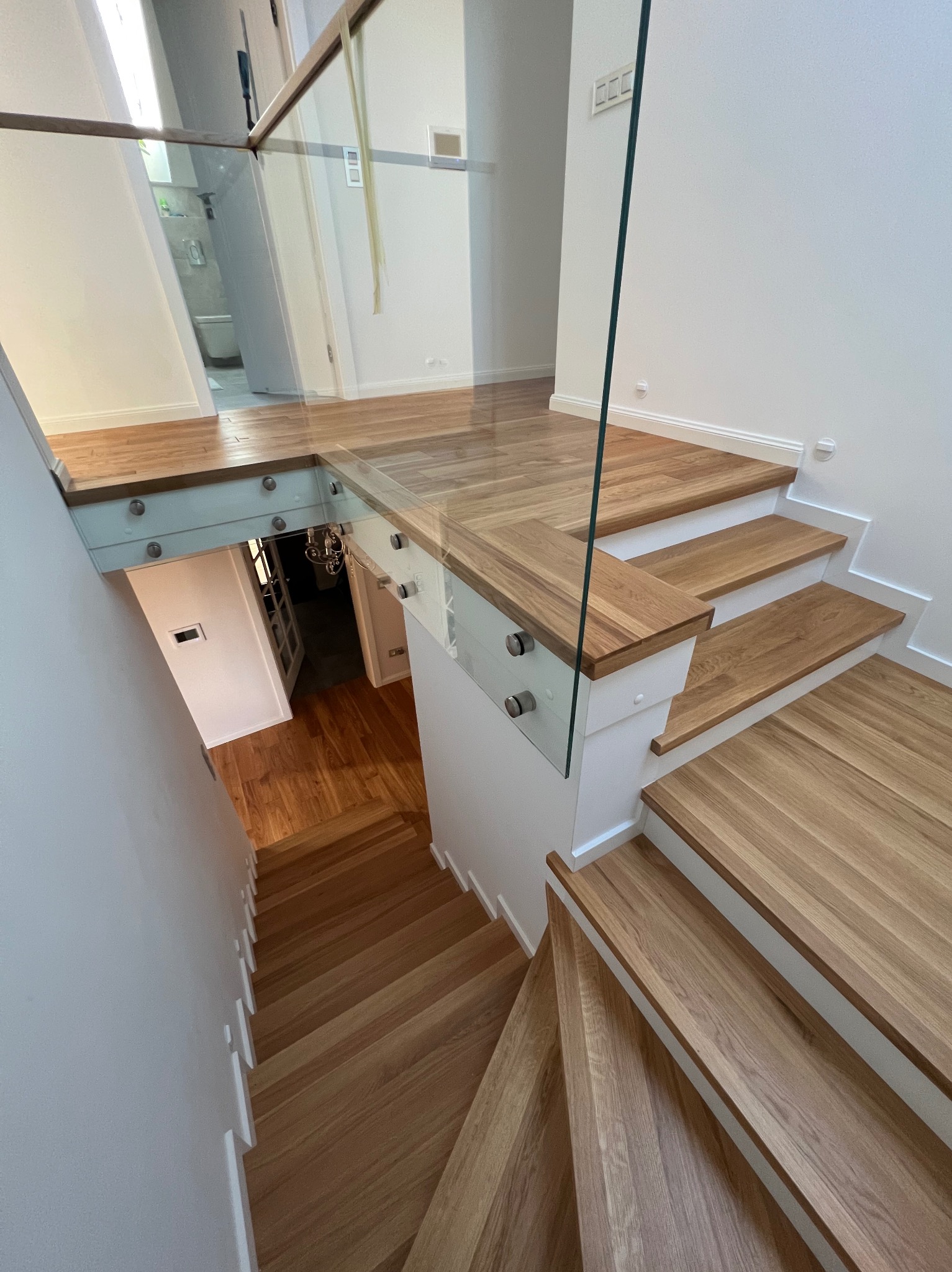 Jasne, drewniane schody z białymi podstopnicami i szklaną balustradą mocowaną na metalowe śruby, widok z góry na dół z widocznym fragmentem podłogi na górnym poziomie.