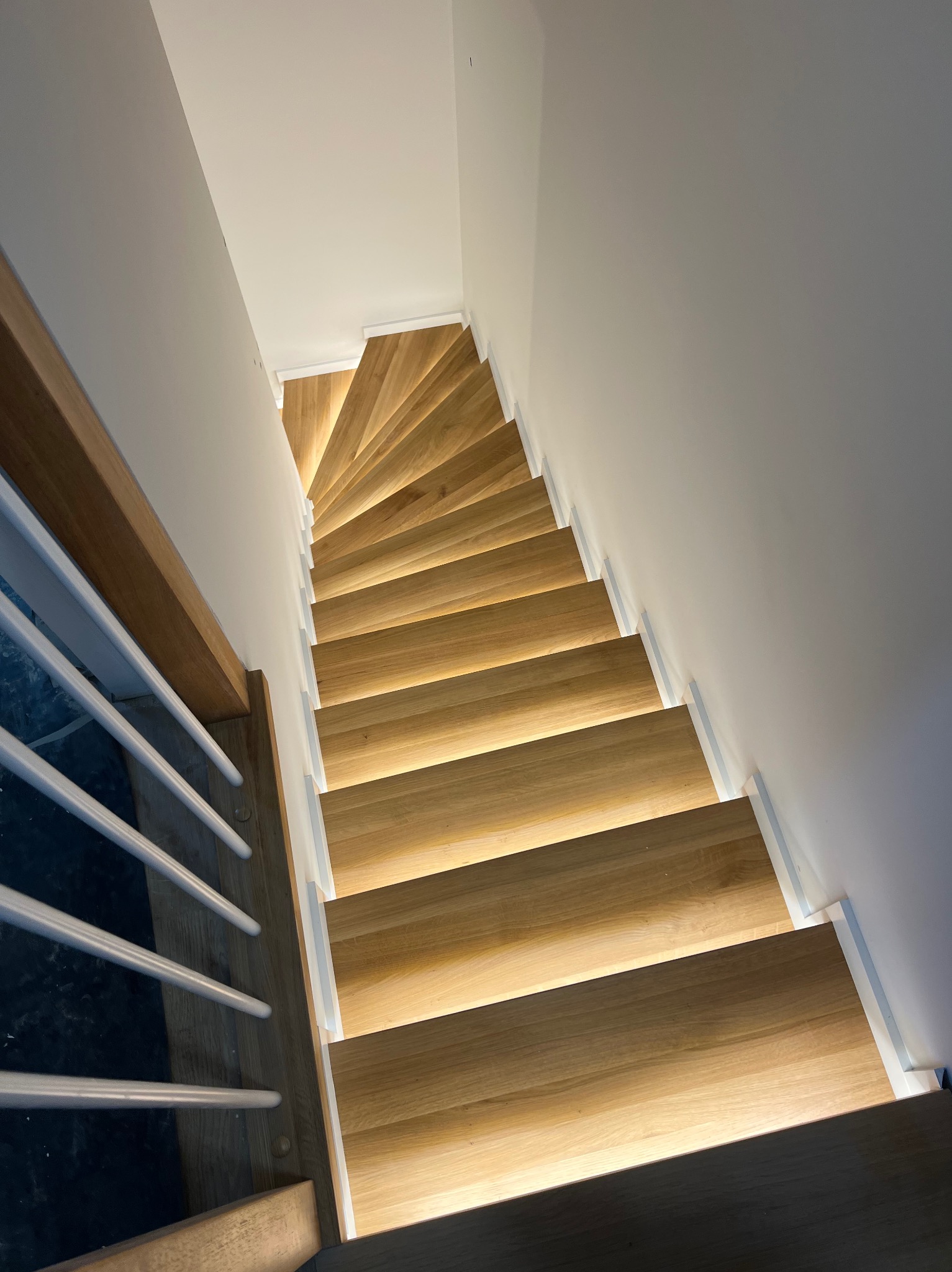 Wysoki kąt widzenia na kręte, jasne drewniane schody z podświetleniem LED wzdłuż stopni, białe ściany i poręcze z metalowymi prętami.