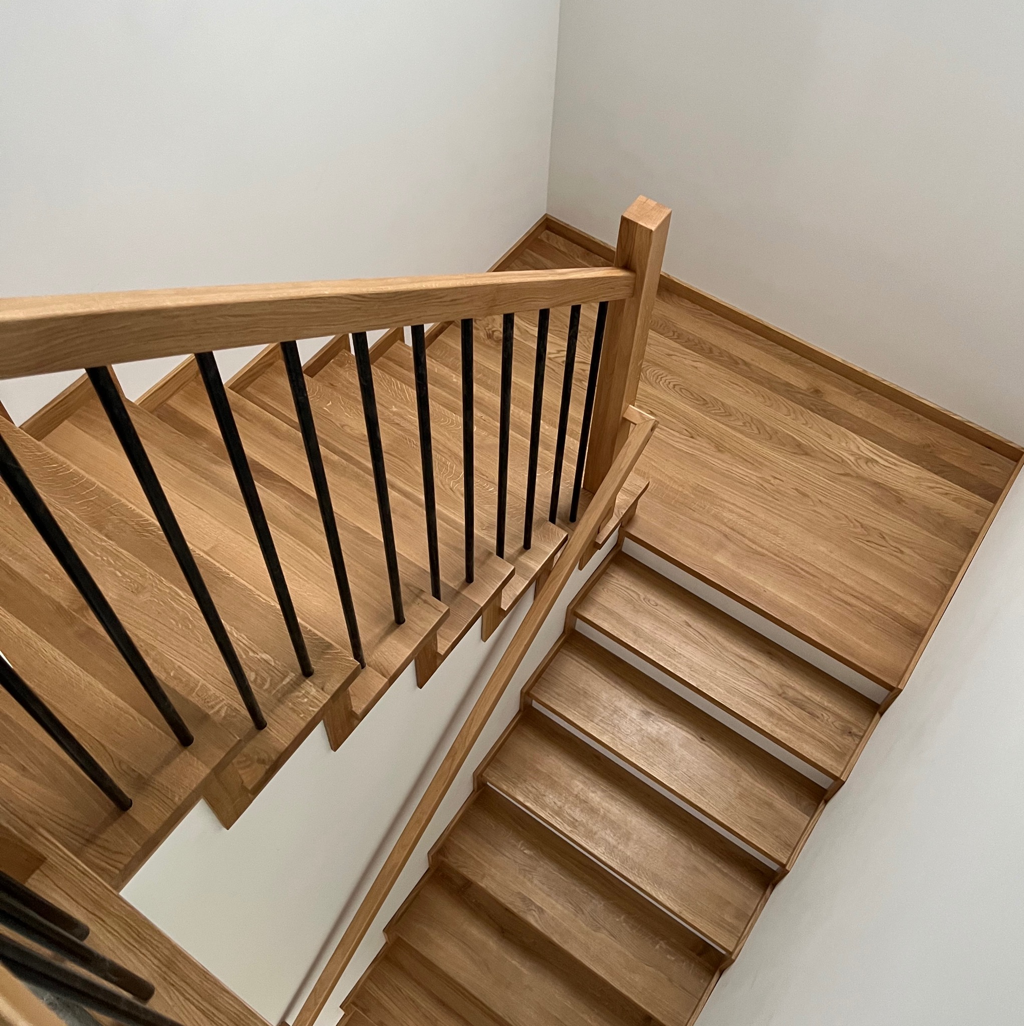 Widok z góry na drewniane schody z czarnymi, metalowymi balustradami w domu w Górze Kalwarii. Naturalne wykończenie drewna kontrastuje z białymi ścianami.