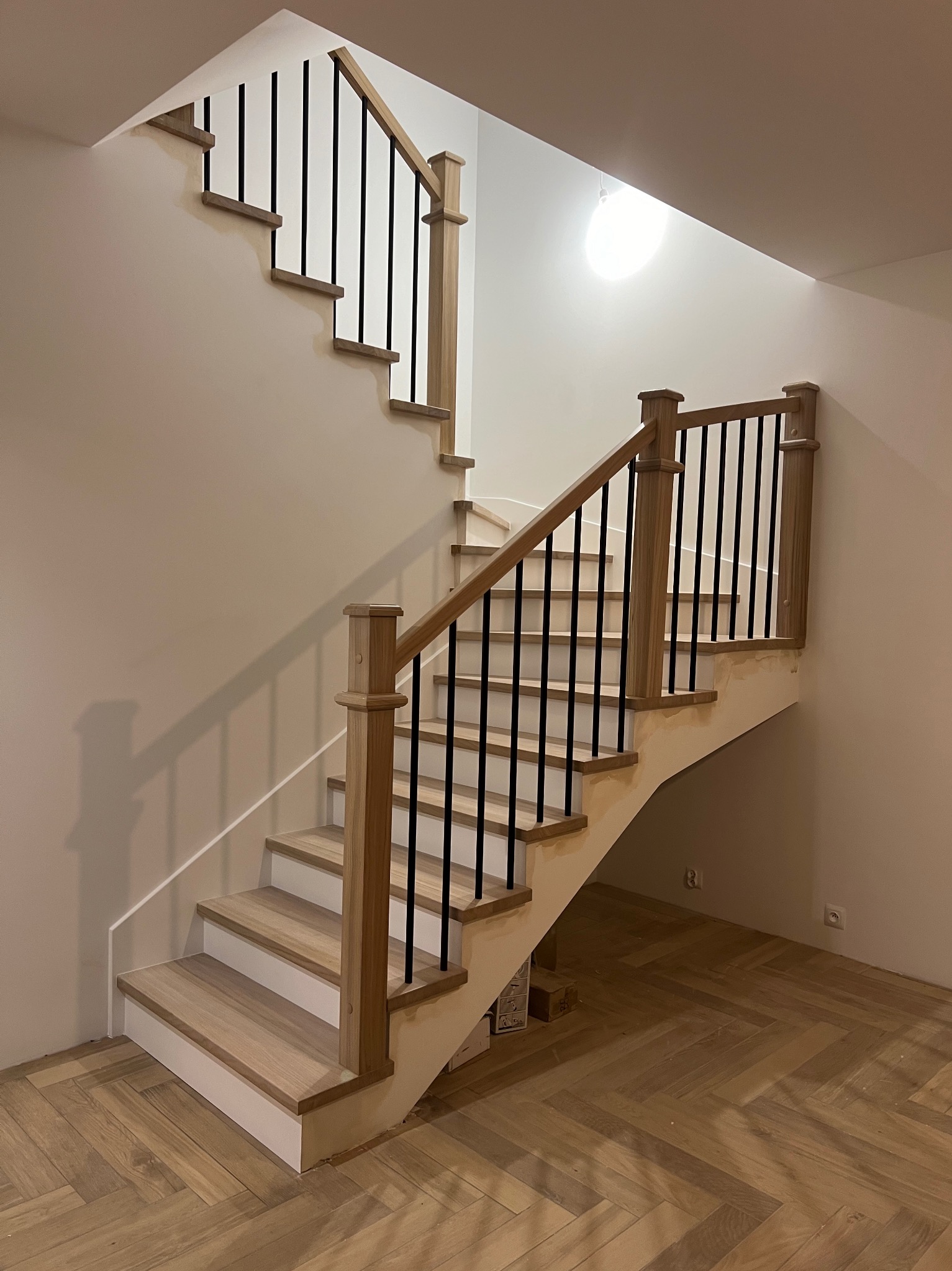 Nowoczesne schody z drewnianymi stopniami i czarnymi balustradami, oświetlone górnym światłem, z parkietem ułożonym w jodełkę na podłodze.