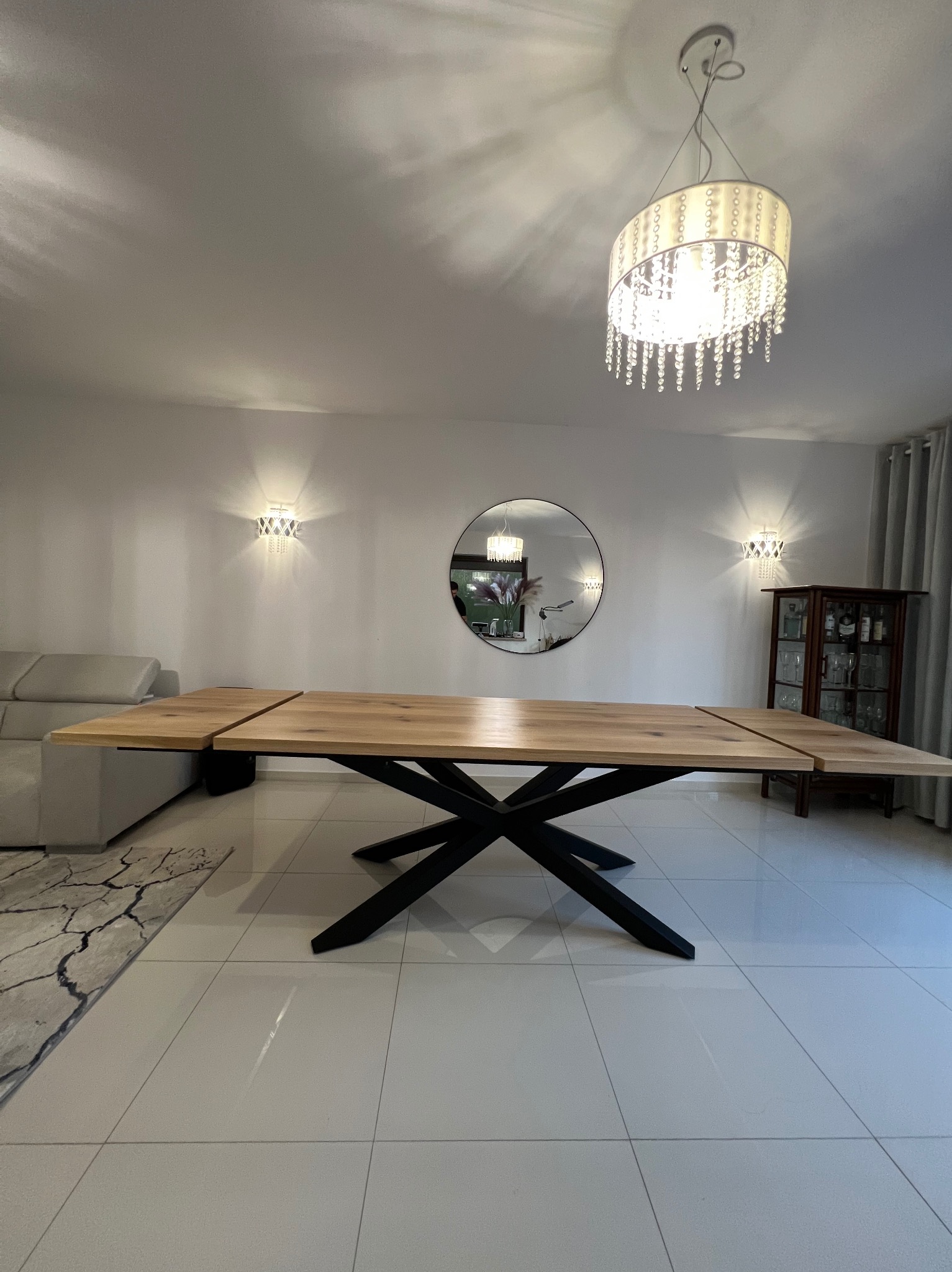 Rozkładany stół z blatem z jasnego drewna i czarną, geometryczną podstawą w nowoczesnym salonie z białymi ścianami, oświetlonym ozdobnym żyrandolem i kinkietami.