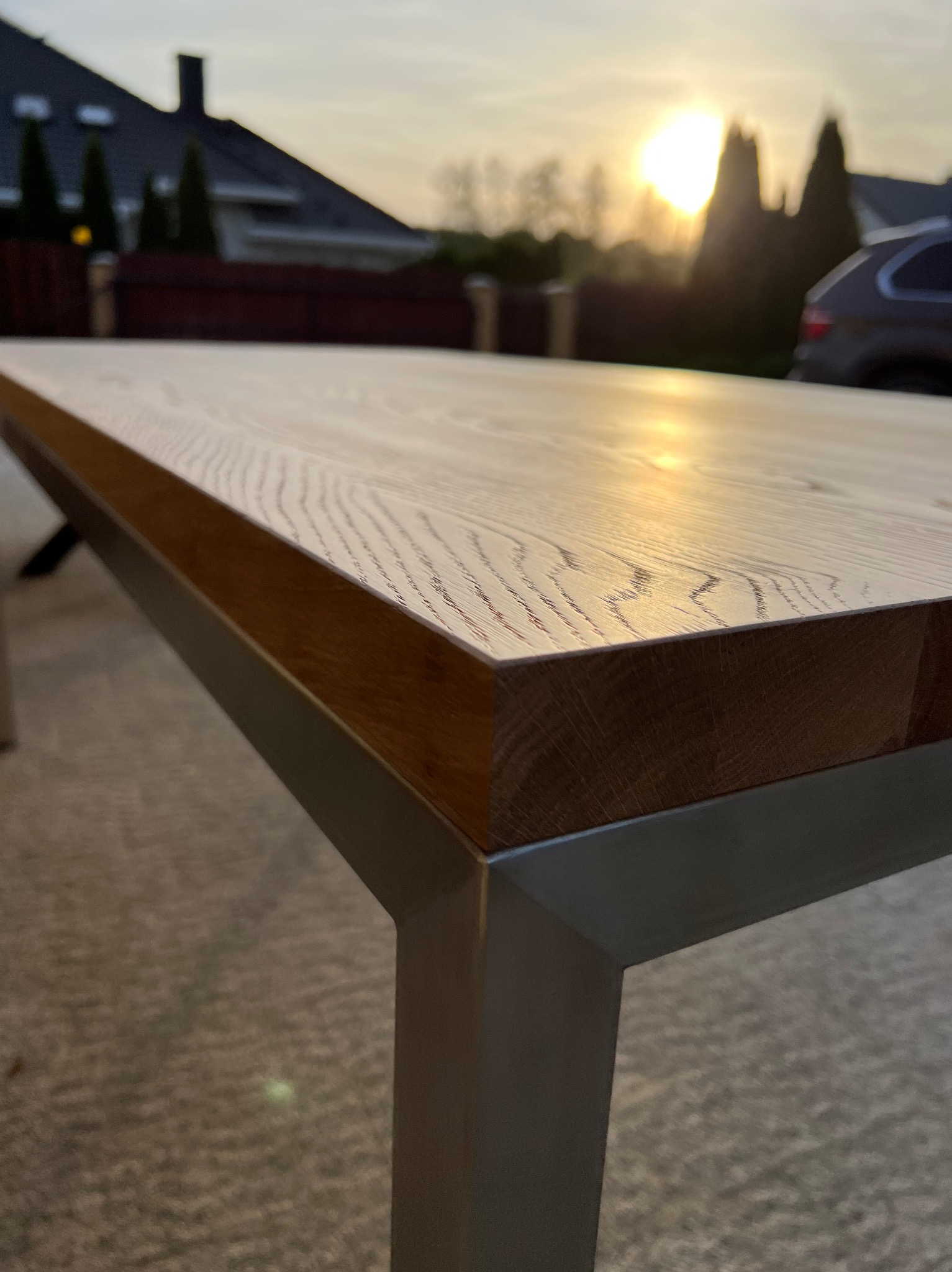Blat drewnianego stołu z metalowymi nogami, sfotografowany na zewnątrz w promieniach zachodzącego słońca, z widocznym domem i samochodem w tle.