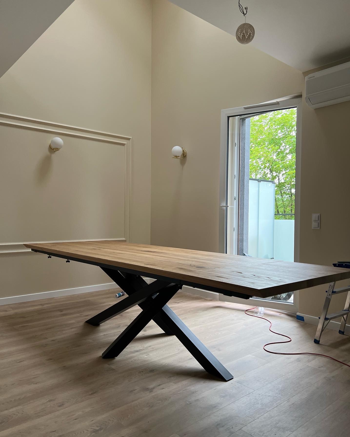 Rozkładany stół z blatem z jasnego drewna i czarnymi, metalowymi nogami w kształcie litery X, ustawiony w jasnym pomieszczeniu z parkietem, widoczne elementy dekoracyjne na ścianach i drabina w tle.