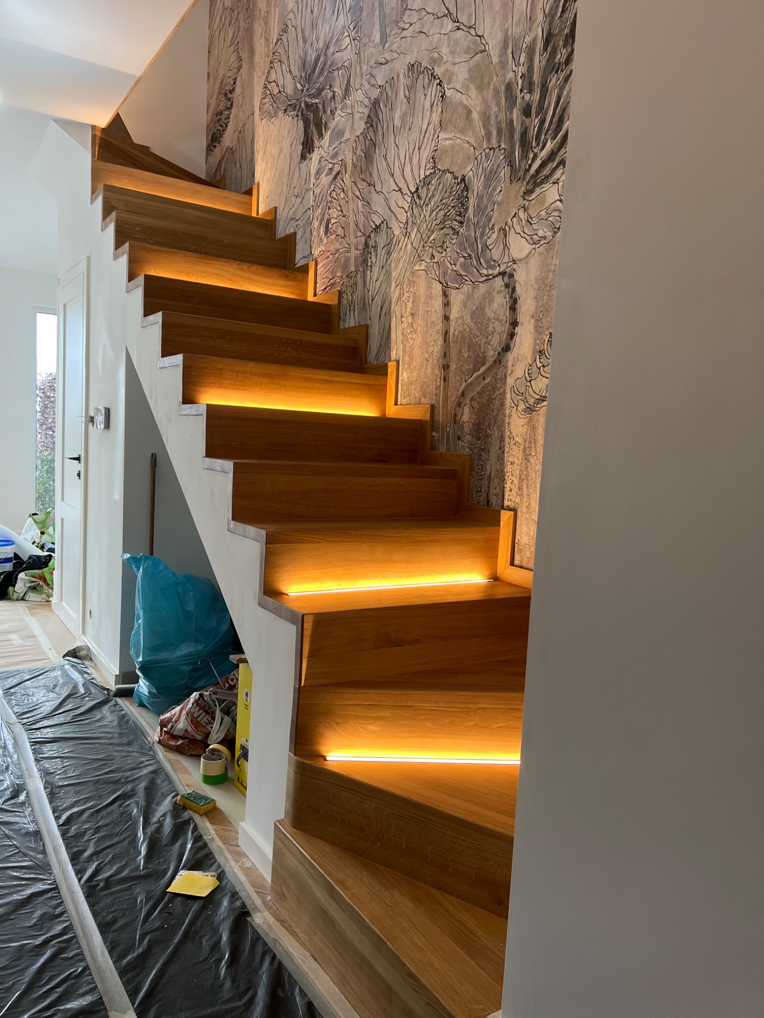 Bieg schodów z drewna z podświetleniem LED na każdym stopniu, widoczne folie ochronne na podłodze i ściana z dekoracyjną tapetą w tle.
