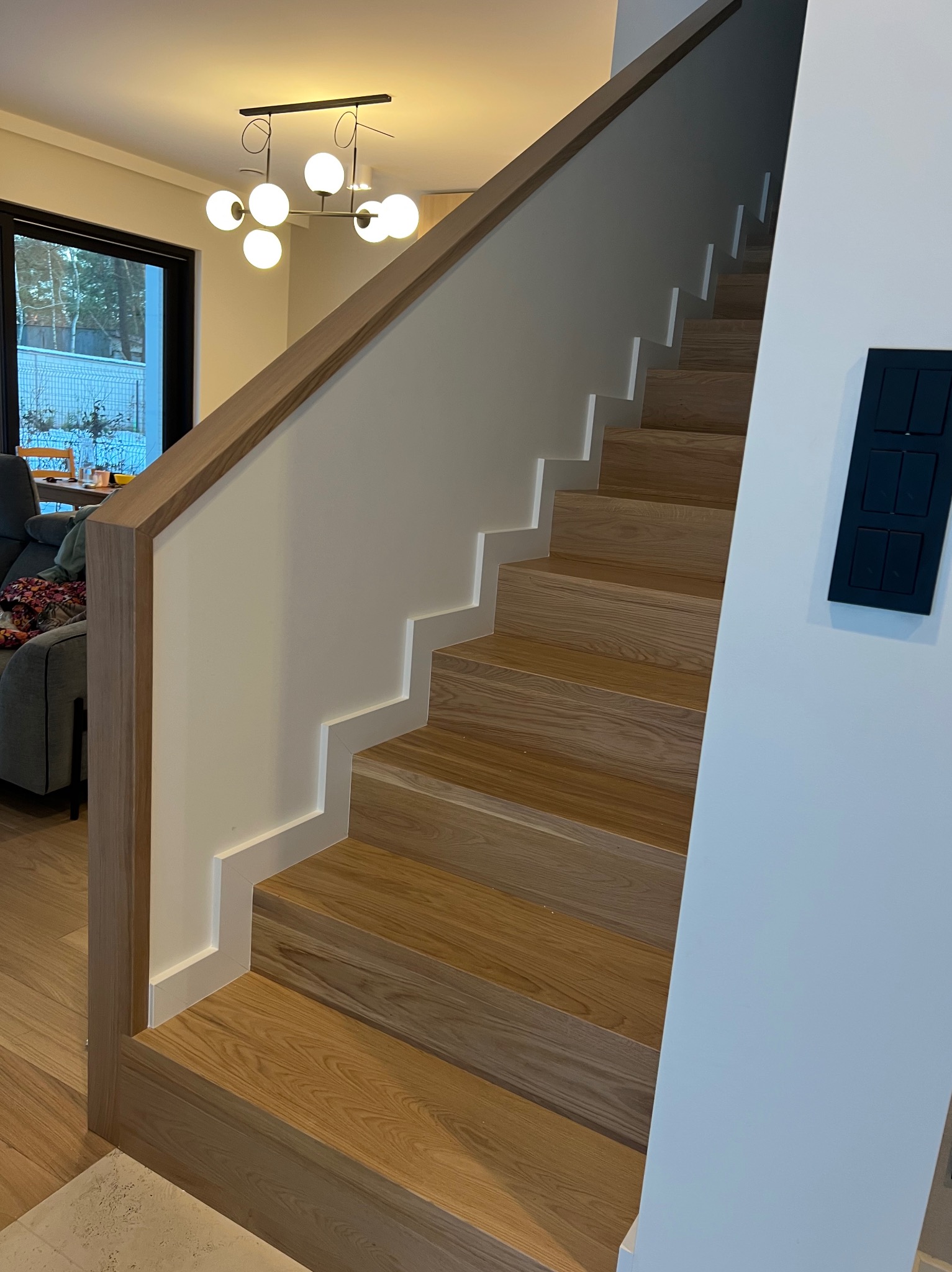 Schody dębowe dywanowe lekko bielone z zabudowaną ścianką, białym cokołem i drewnianym pochwytem w kolorze schodów.