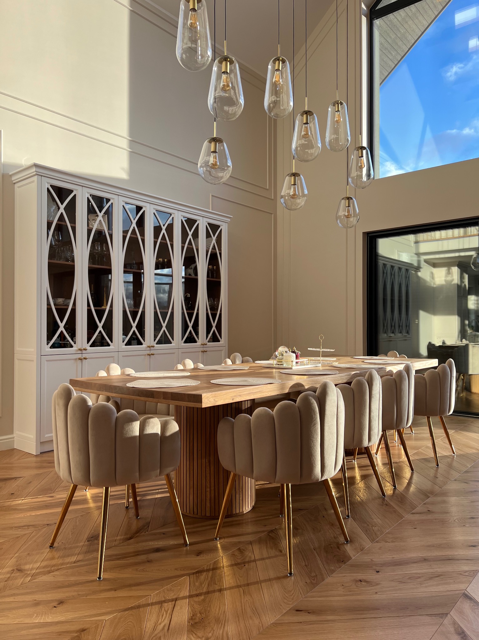 Jadalnia z dębowym stołem na okrągłej ryflowanej podstawie, otoczonym beżowymi krzesłami na złotych nogach, na parkiecie ułożonym w jodełkę, z białym kredensem w tle i designerskimi lampami wiszącymi.