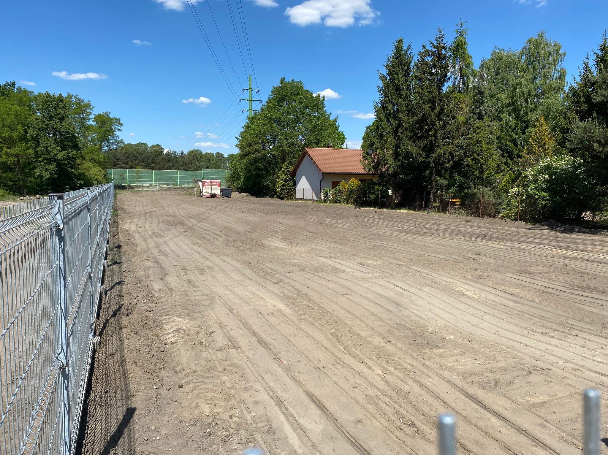 Przygotowany teren pod budowę, widoczne ślady po maszynach, ogrodzenie z siatki, w tle budynek mieszkalny i słup wysokiego napięcia.