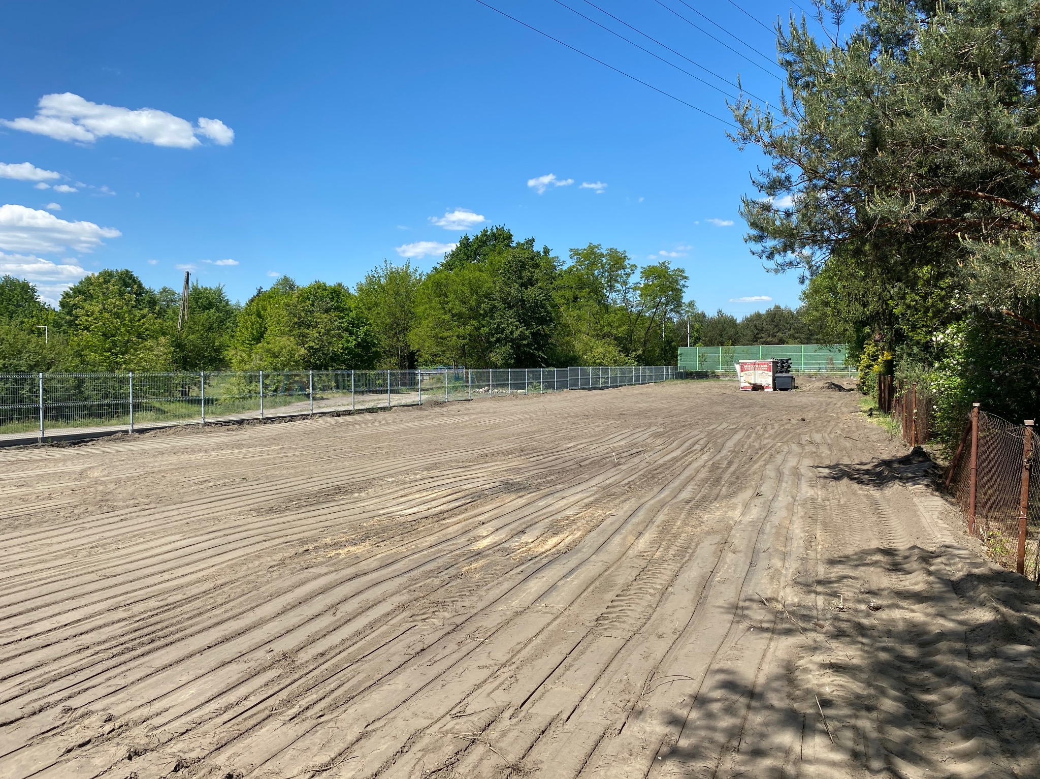 Wyrównany teren budowlany z widocznymi śladami opon, ogrodzony siatką, z drzewami w tle i błękitnym niebem.