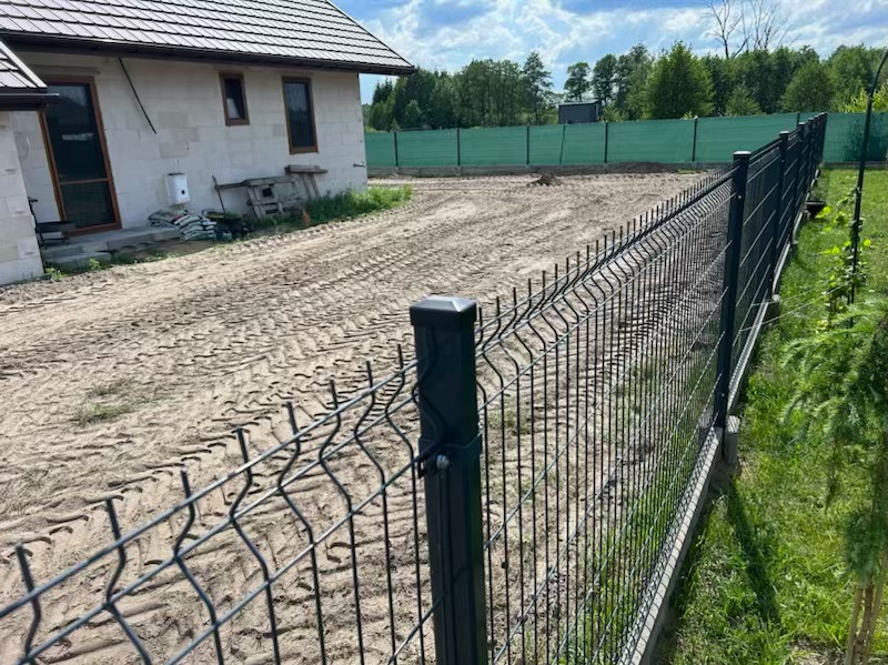 Nowo postawione, panelowe ogrodzenie w kolorze grafitowym na działce z wyrównanym terenem, widoczne ślady opon i budynek w stanie surowym z workami cementu w tle.
