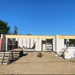 Wznoszenie ścian parterowego domu z bloczków betonu komórkowego, widoczne stemple podtrzymujące nadproża, przygotowane materiały budowlane i drewniane schodki na placu budowy w słoneczny dzień.