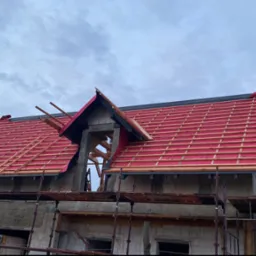 Budowa domu: widok na dach w trakcie krycia z widoczną membraną w kolorze czerwonym i drewnianą konstrukcją łat, rusztowanie w tle.