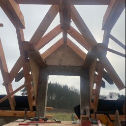 Ihor Kadynskyi - Konstrukcja drewniana dachu w trakcie budowy, widoczne krokwie i deski, narzędzia na prowizorycznym rusztowaniu, w tle las i pochmurne niebo.